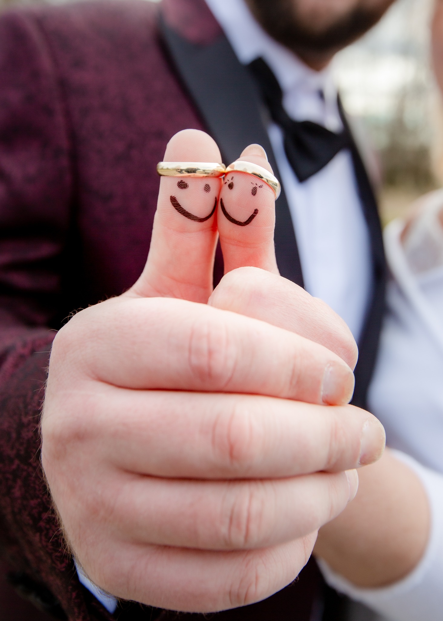 Dwa kciuki z narysowanymi uśmiechniętymi buziami i złotymi obrączkami, na tle marynarki i muchy pana młodego oraz sukni panny młodej.