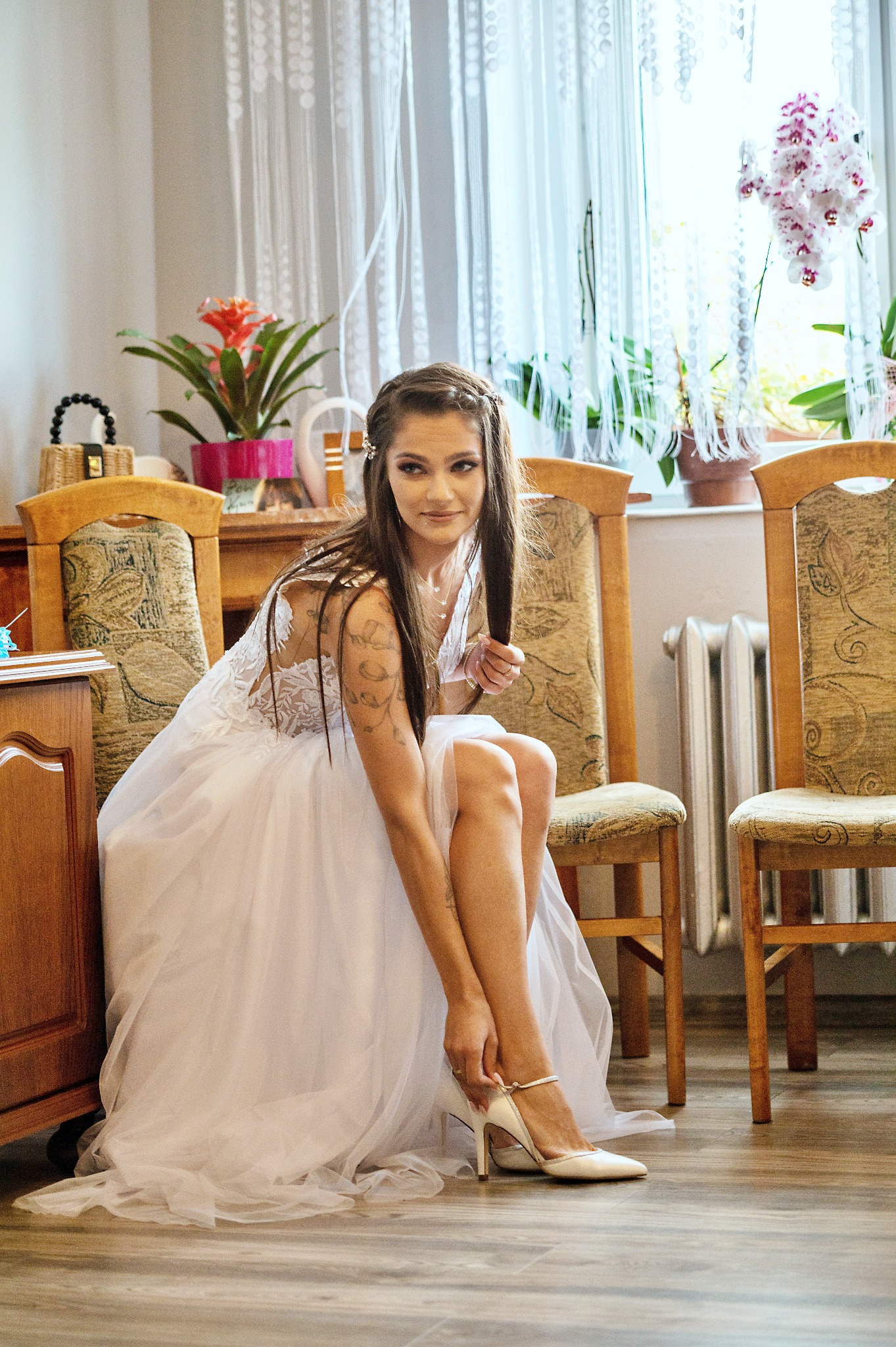 Panna młoda w białej sukni, siedząc na krześle, zapina biały but na obcasie, widoczne kwiaty w doniczkach w tle.