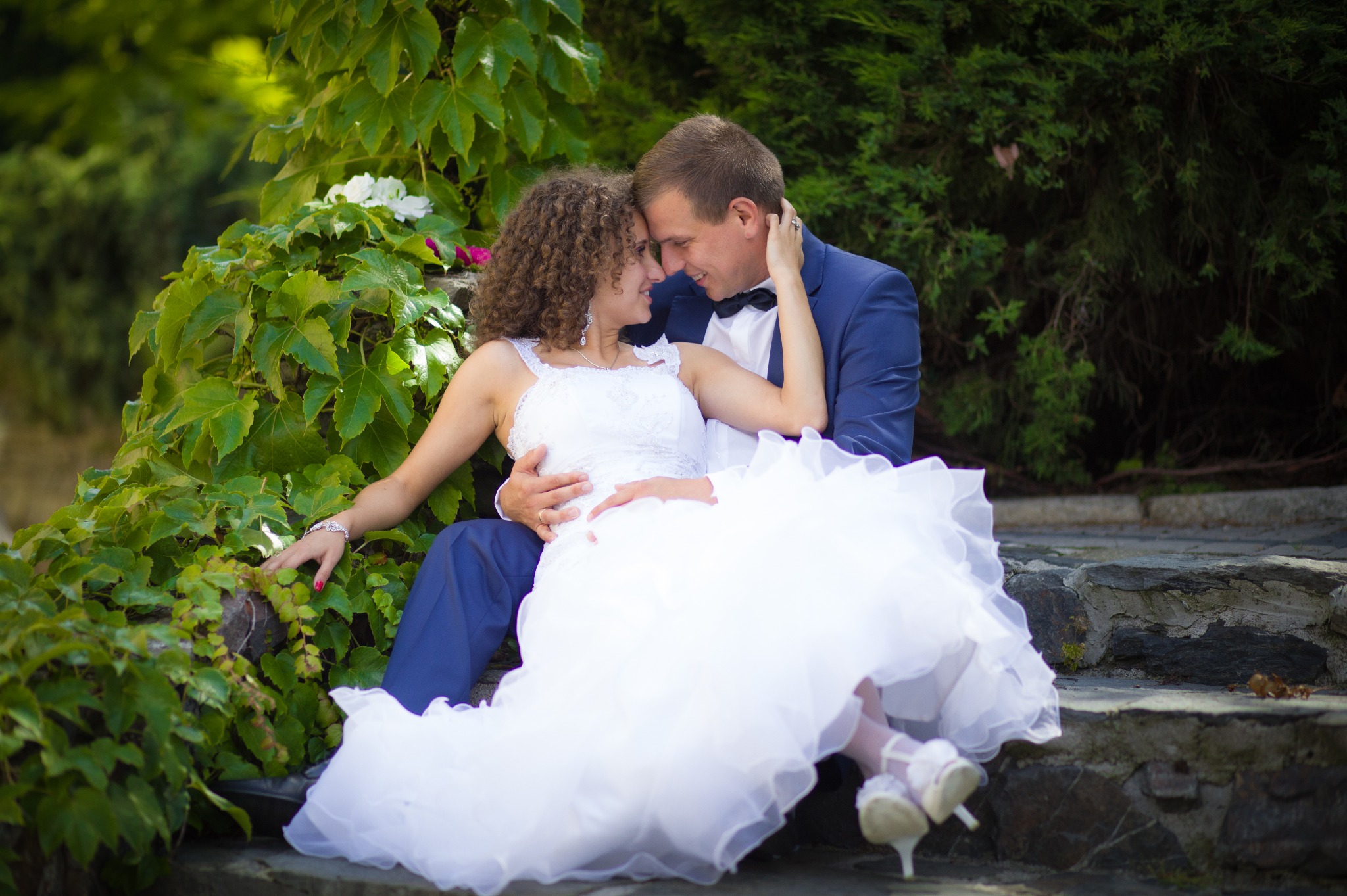 Para młoda siedzi na kamiennych schodach, oparta o ścianę porośniętą bluszczem. Panna młoda w białej sukni z falbanami i ozdobnymi butami, pan młody w granatowym garniturze z muchą. Oboje patrzą...