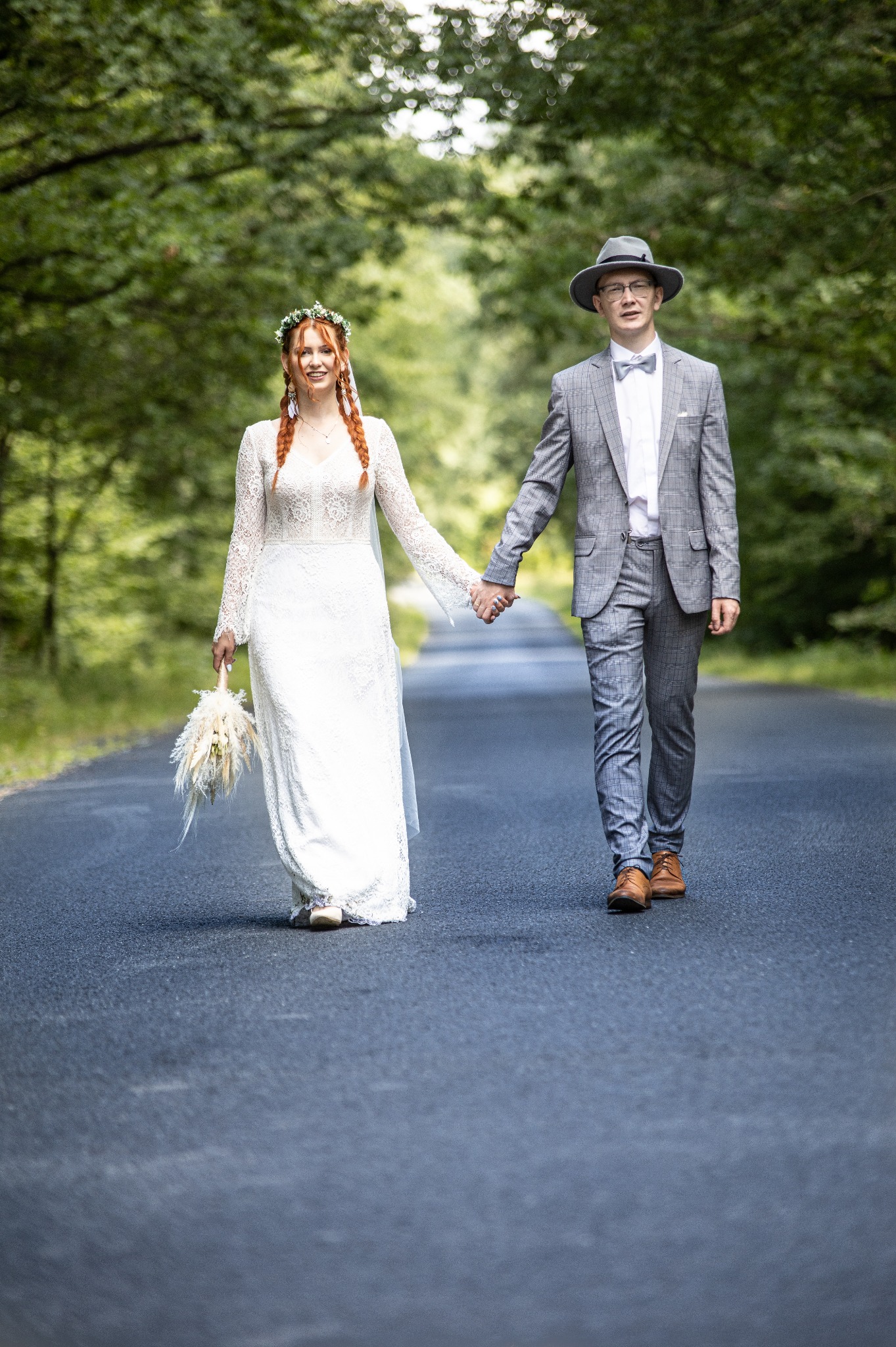 Para młoda, on w szarym garniturze w kratę i kapeluszu, ona w koronkowej sukni z wiankiem na głowie i rudymi warkoczami, trzymają się za ręce idąc asfaltową drogą otoczoną drzewami.