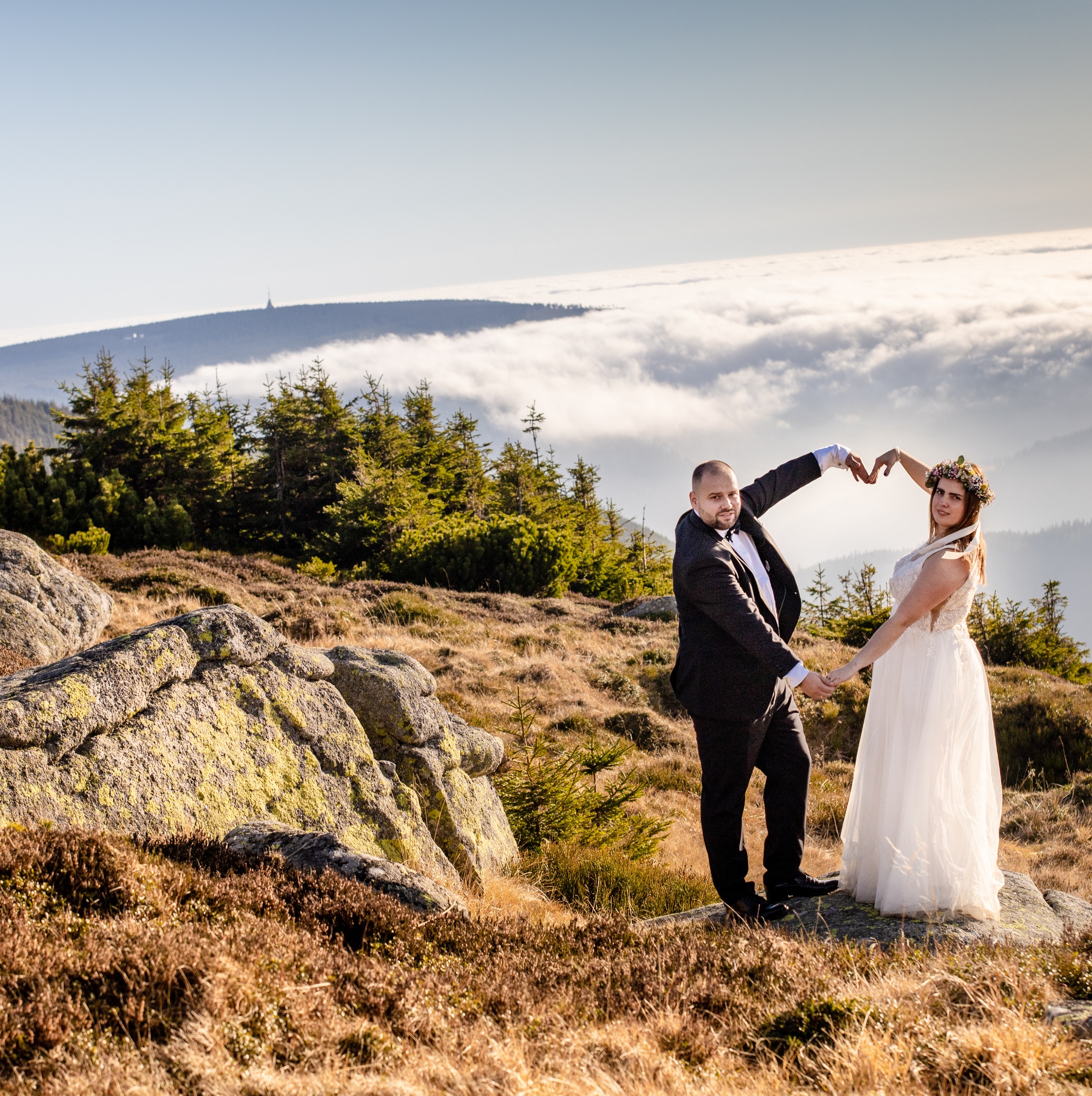 Para młoda w strojach ślubnych stoi na tle górskiego krajobrazu z chmurami, tworząc rękami kształt serca.