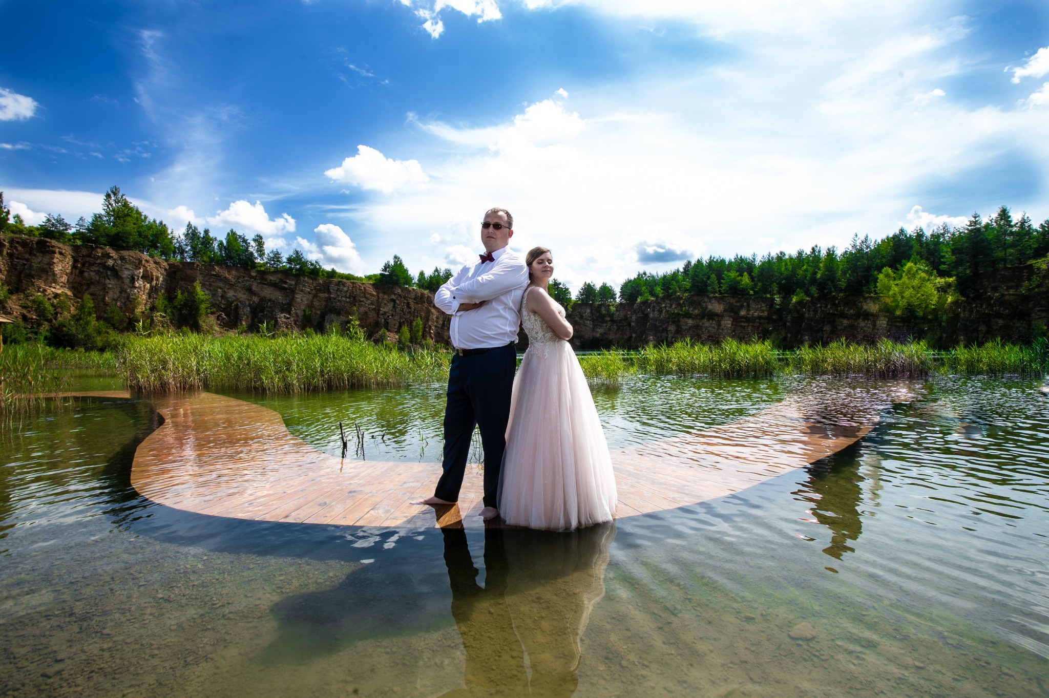 Para w strojach ślubnych stoi na drewnianym pomoście zanurzonym w wodzie, tło stanowi skalisty brzeg porośnięty zielenią i błękitne niebo.