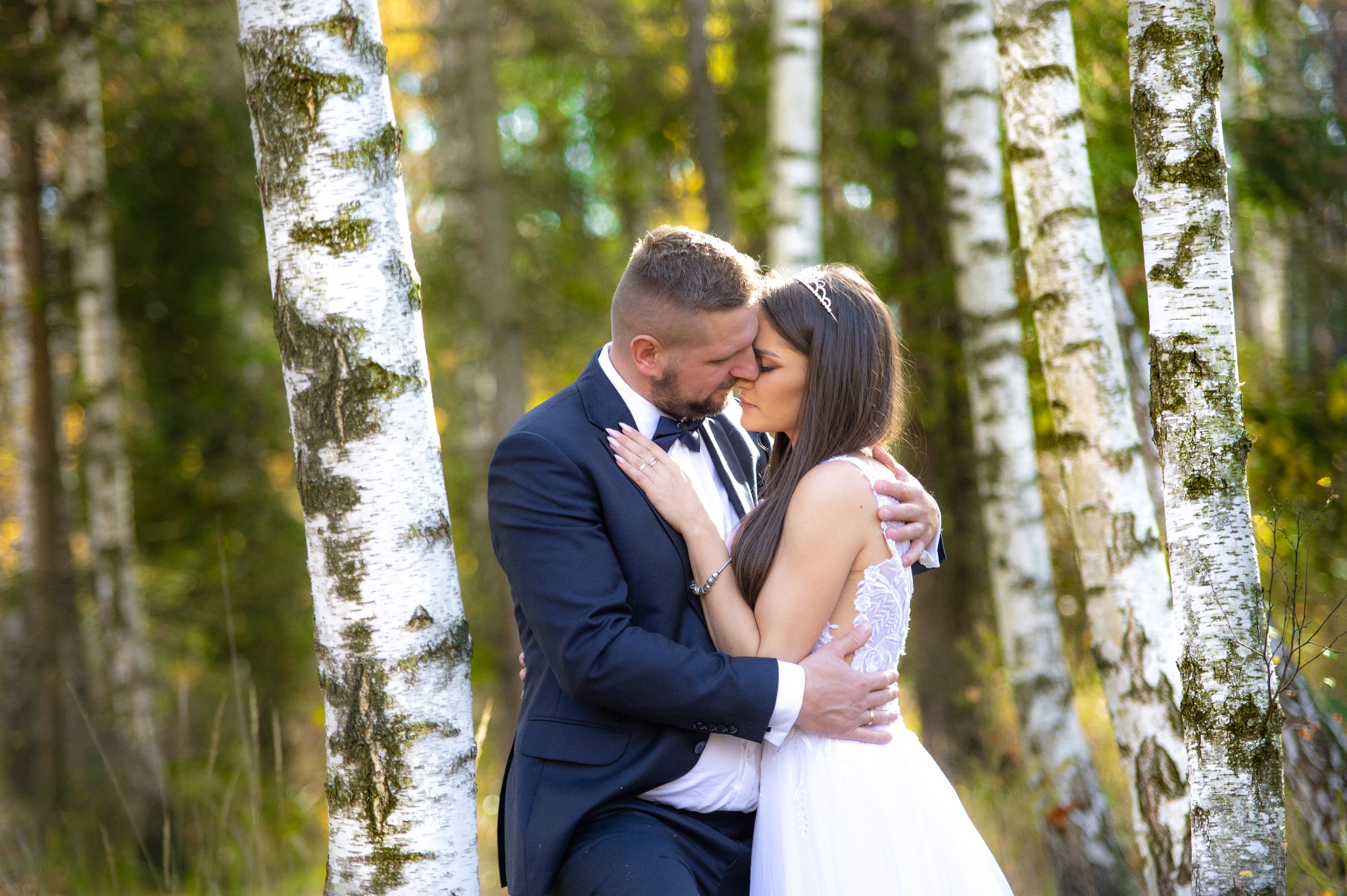 Para młoda w objęciach na tle brzozowego lasu, panna młoda w koronie i sukni z koronkowymi plecami, pan młody w granatowym garniturze z muchą.