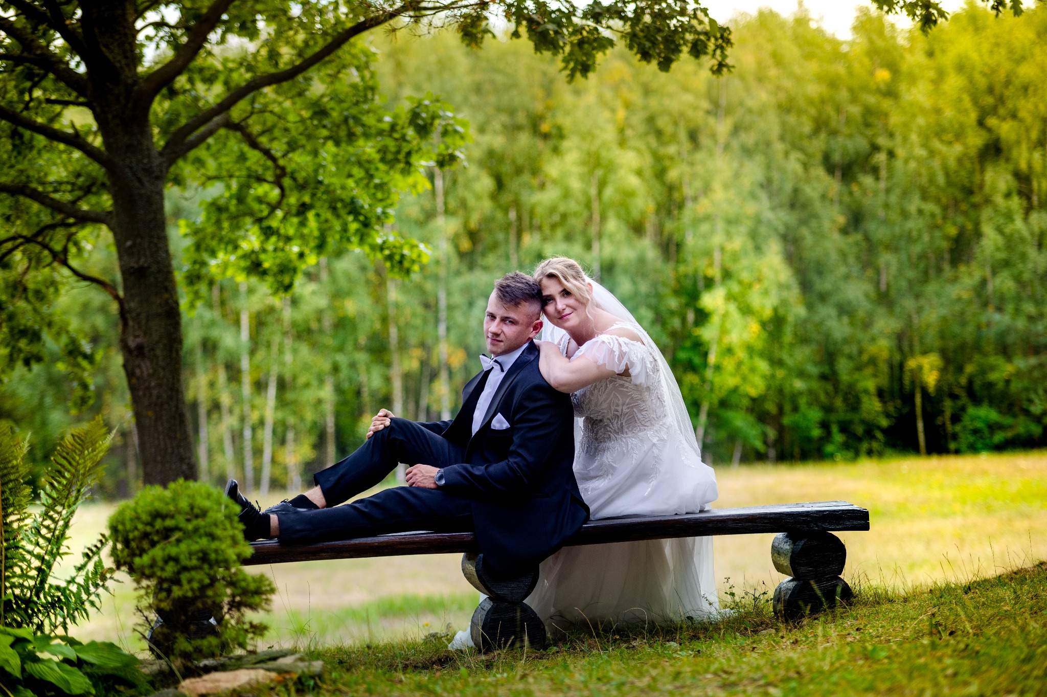 Para młoda w strojach ślubnych siedzi na drewnianej ławce w otoczeniu zieleni, panna młoda opiera głowę na ramieniu pana młodego, w tle drzewa.