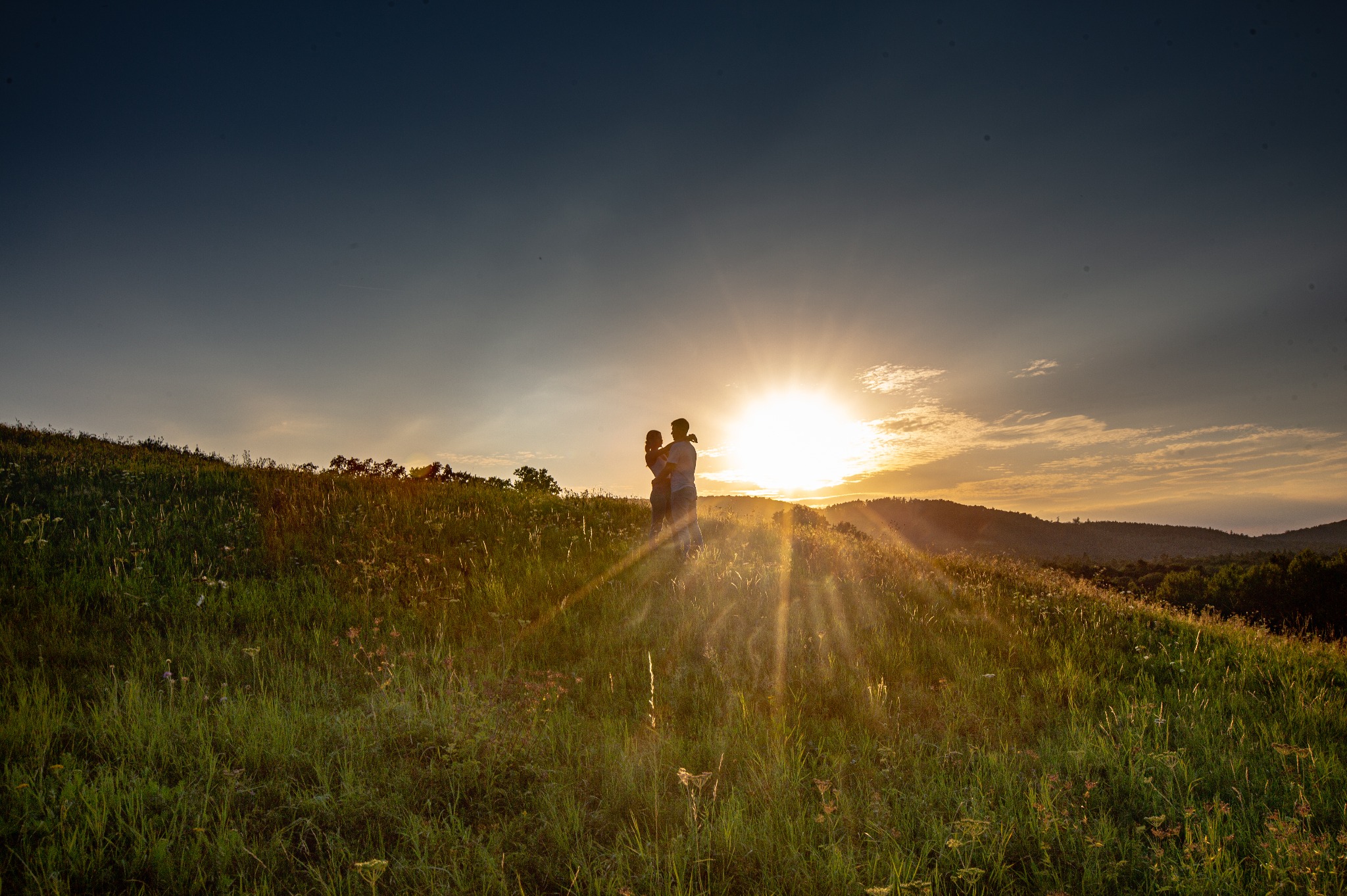 Para zakochanych obejmująca się na szczycie zielonego wzgórza podczas zachodu słońca, z promieniami słonecznymi przebijającymi się przez trawę.