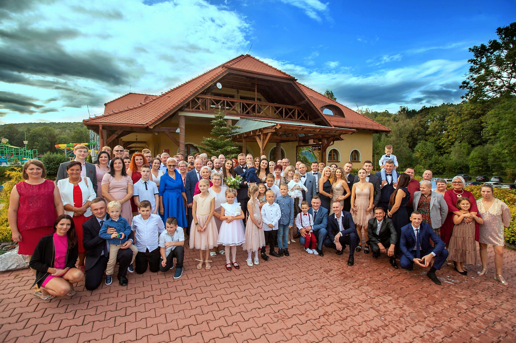 Duża grupa ludzi, w tym dzieci, pozuje do zdjęcia na zewnątrz budynku z brązowym dachem i czerwonej kostki brukowej. Niebo jest częściowo zachmurzone.