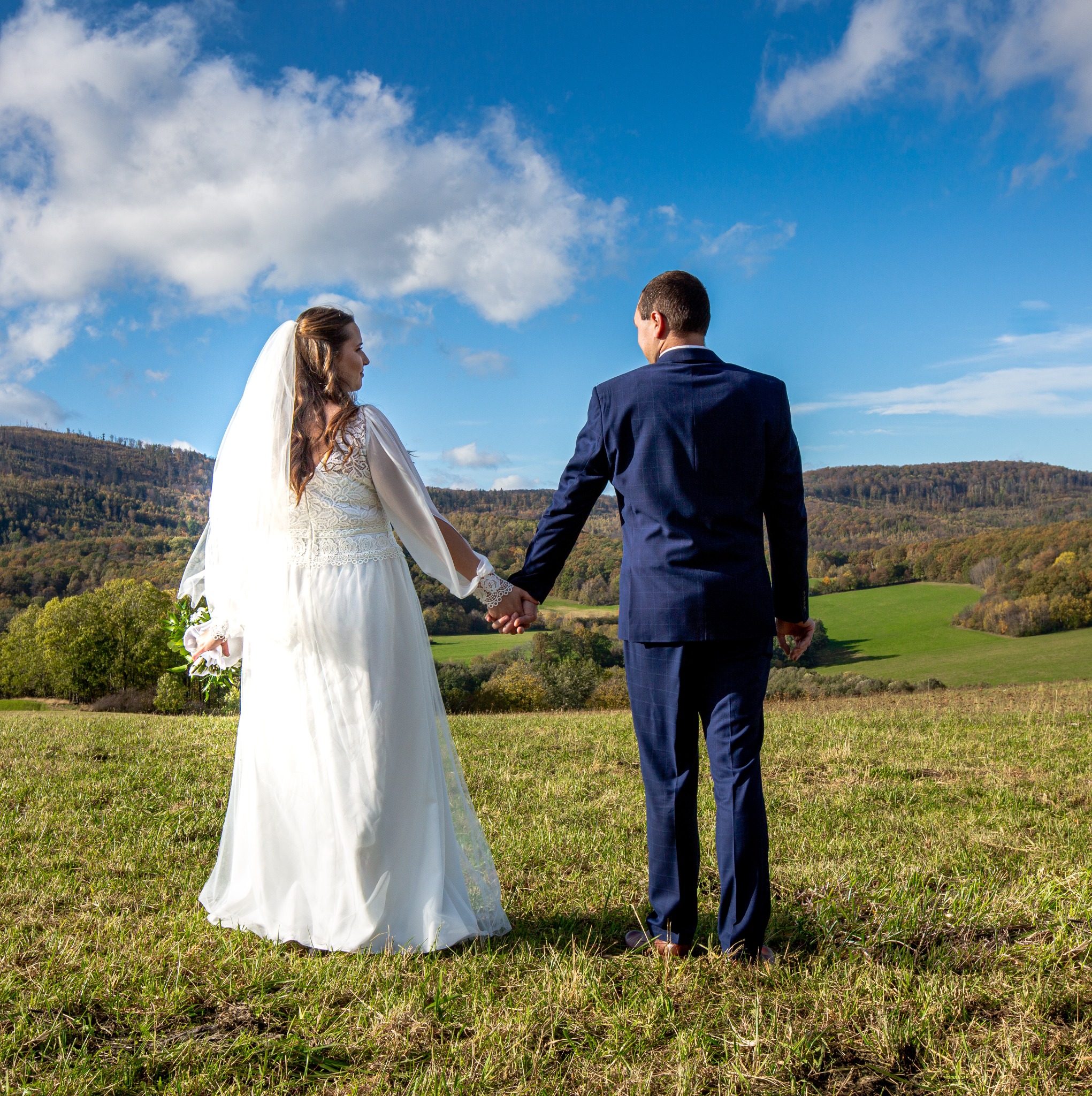 Para młoda trzymająca się za ręce, odwrócona tyłem, w tle zielone wzgórza i błękitne niebo z chmurami. Panna młoda w białej sukni z koronkowymi rękawami i welonem, pan młody w granatowym garniturze...
