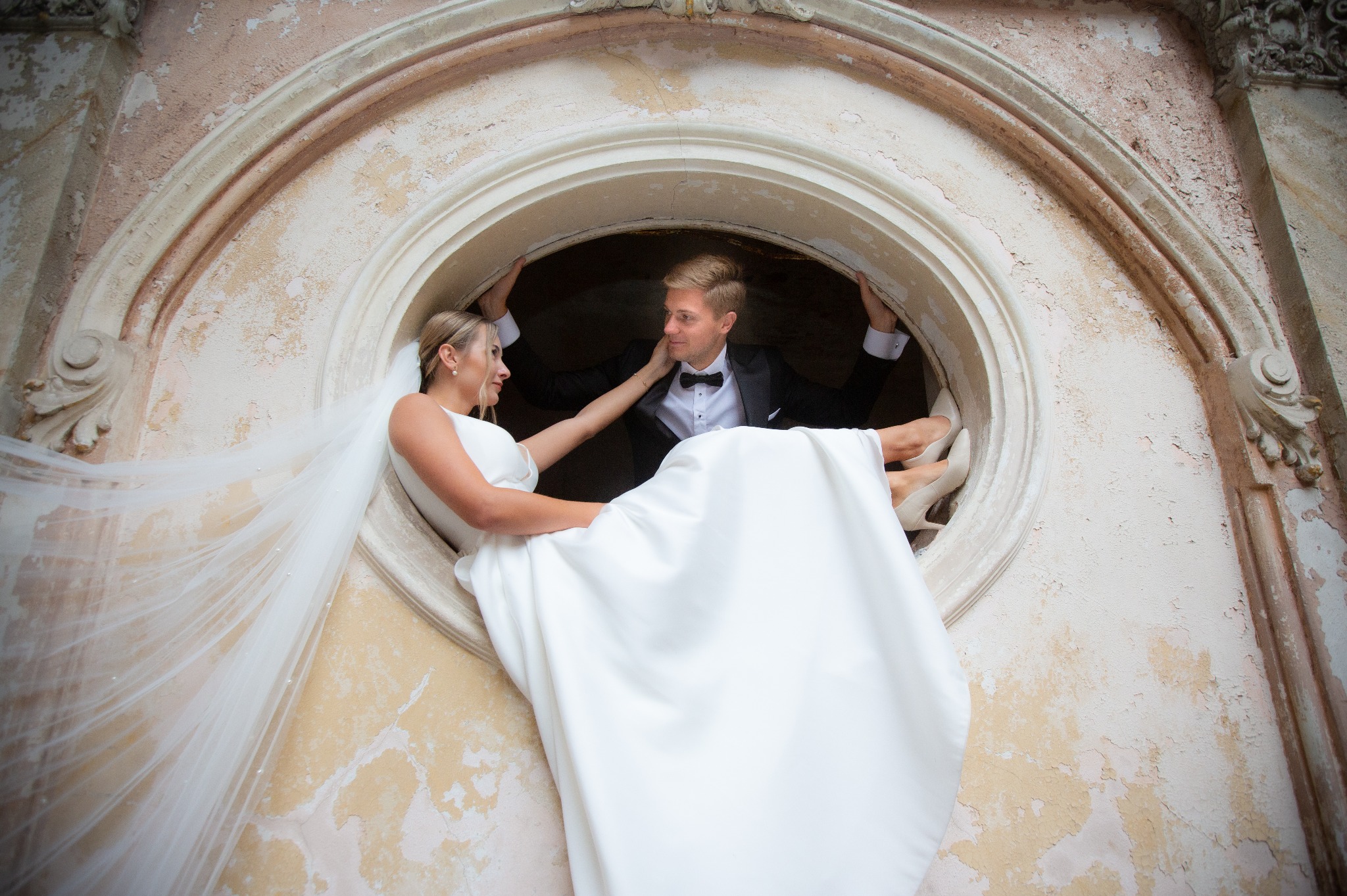 Para młoda w oknie o okrągłym kształcie: panna młoda w białej sukni i welonie opiera się o parapet, pan młody w czarnym smokingu trzyma się ramy okna, spoglądając na nią z uśmiechem.