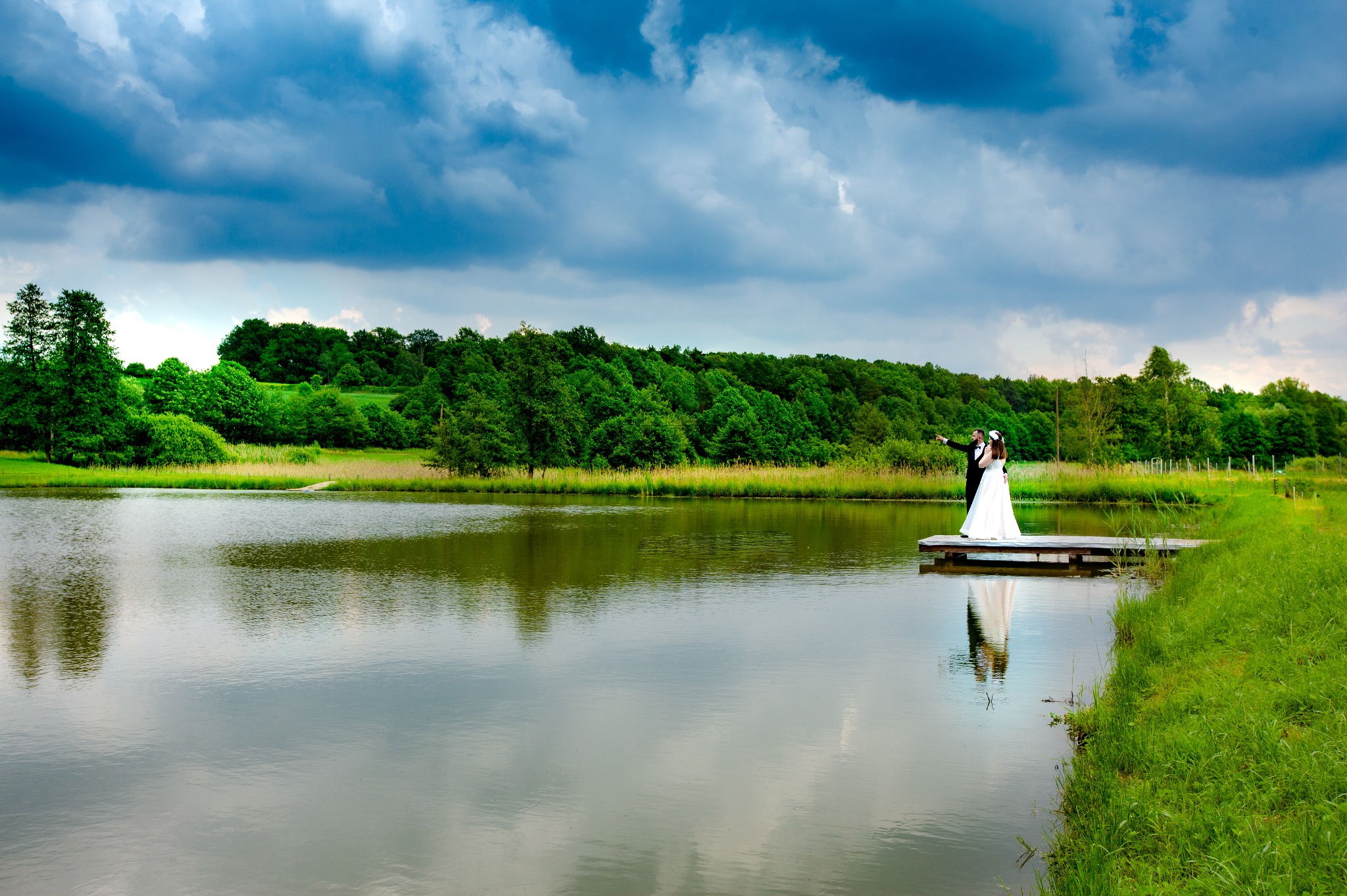 Para młoda w strojach ślubnych stoi na drewnianym pomoście nad jeziorem, zalesiony brzeg i dramatyczne chmury w tle.
