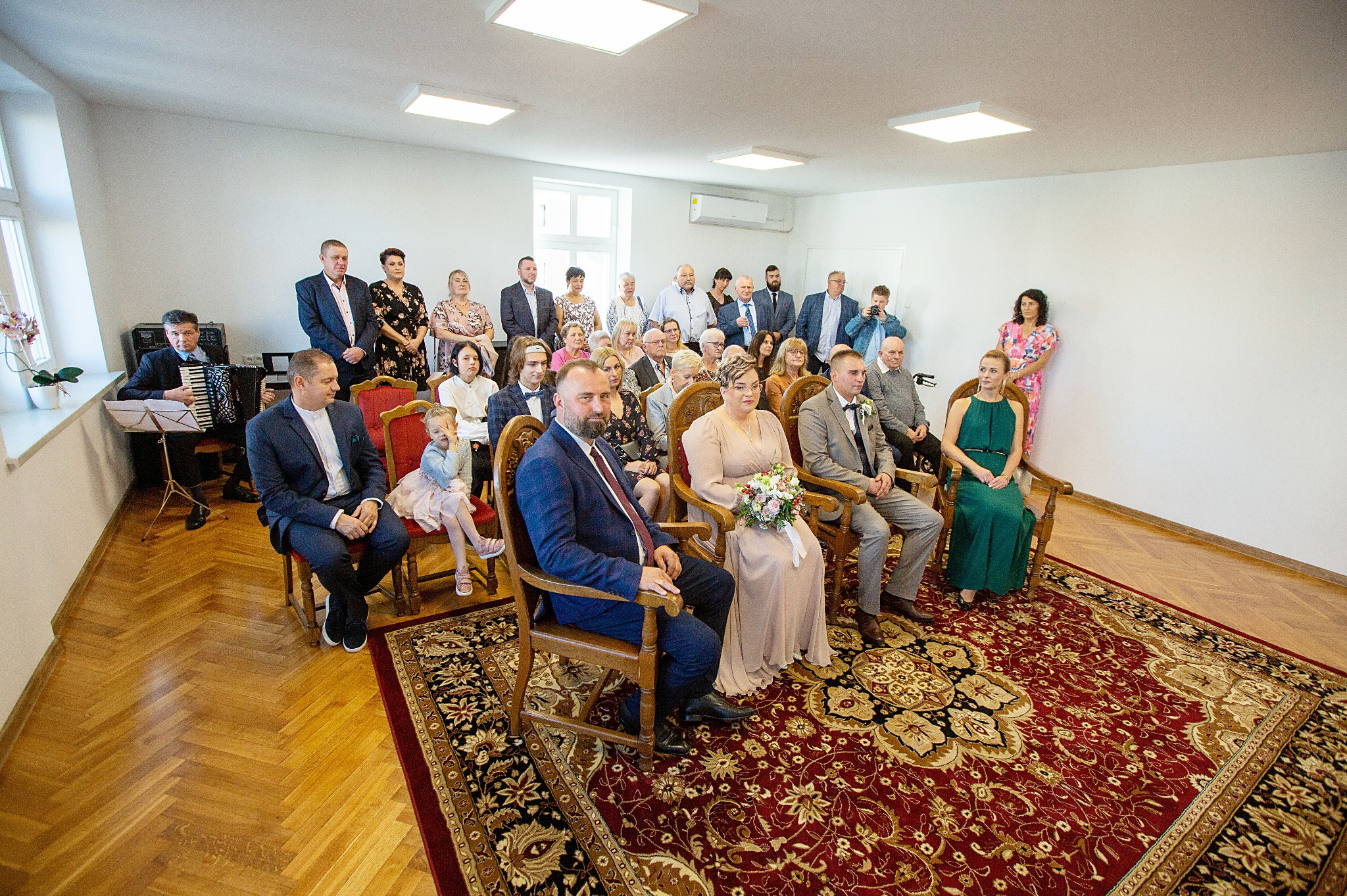 Sala USC podczas ceremonii ślubnej: para młoda siedzi na krzesłach, w tle goście, akordeonista i czerwony dywan z wzorami.