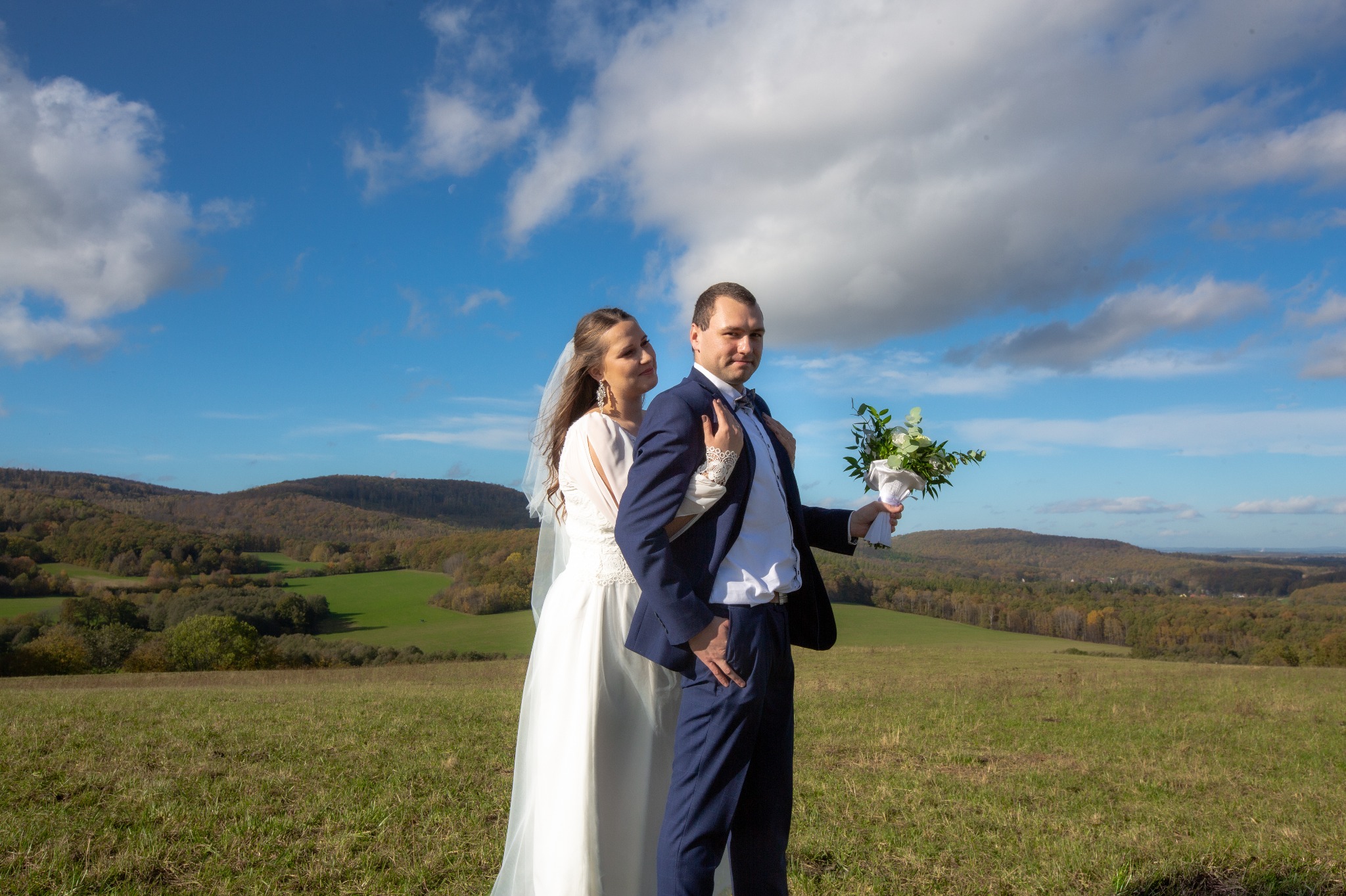 Para młoda w plenerze na tle pagórkowatego krajobrazu i błękitnego nieba z chmurami. Panna młoda w białej sukni z welonem obejmuje pana młodego w granatowym garniturze, który trzyma bukiet.