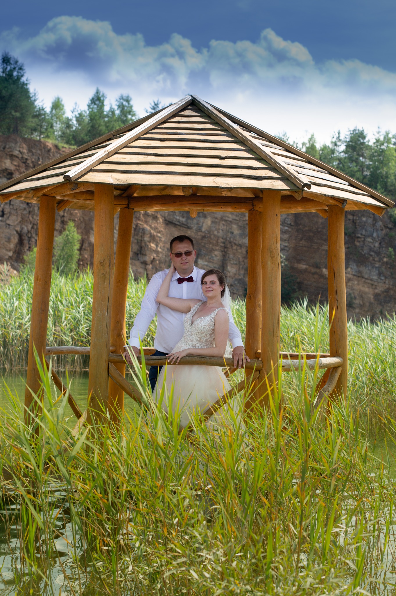 Para młoda w drewnianej altance nad jeziorem, panna młoda w sukni ecru z koronkową górą, pan młody w białej koszuli i bordowej muszce, trzciny wodne w pierwszym planie, skaliste wzgórze w tle.