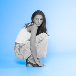 Helena Piecuch - Kobieta w białym topie i jeansach w kucki, z rękami na stopie w czarnym szpilce, na jednolitym błękitnym tle.