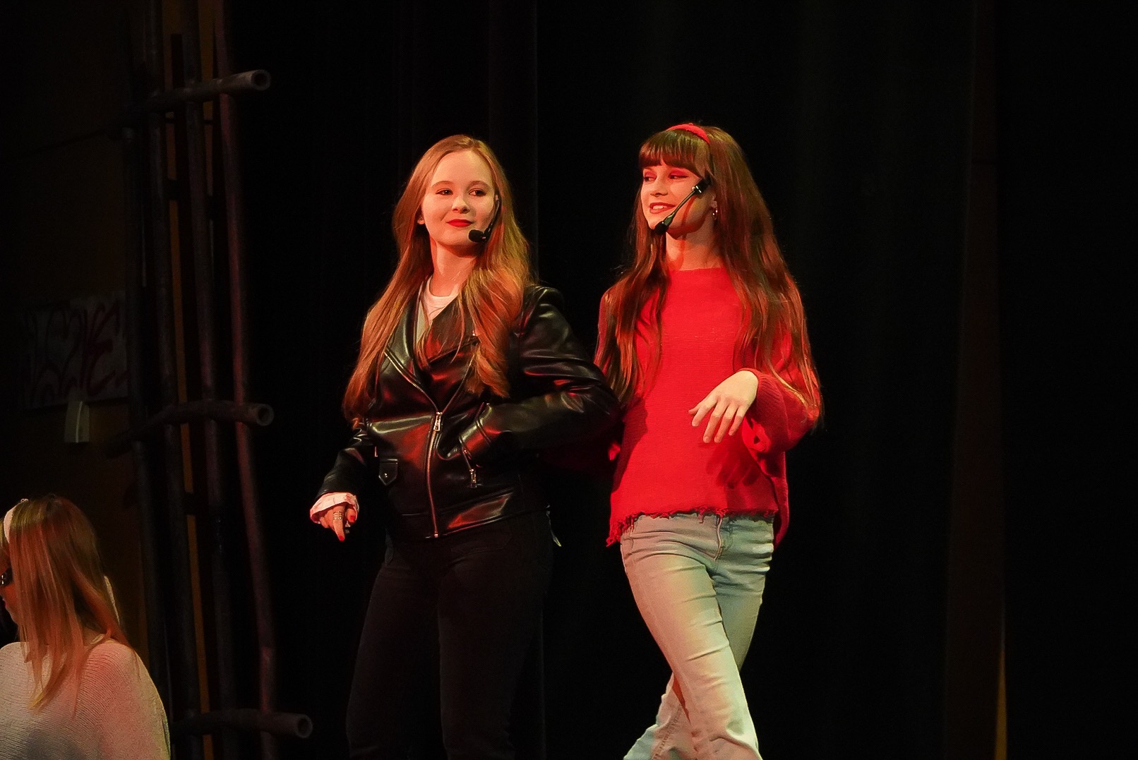 Dwie młode kobiety z mikroportami na scenie, jedna w czarnej skórzanej kurtce, druga w czerwonym swetrze, oświetlenie sceniczne.