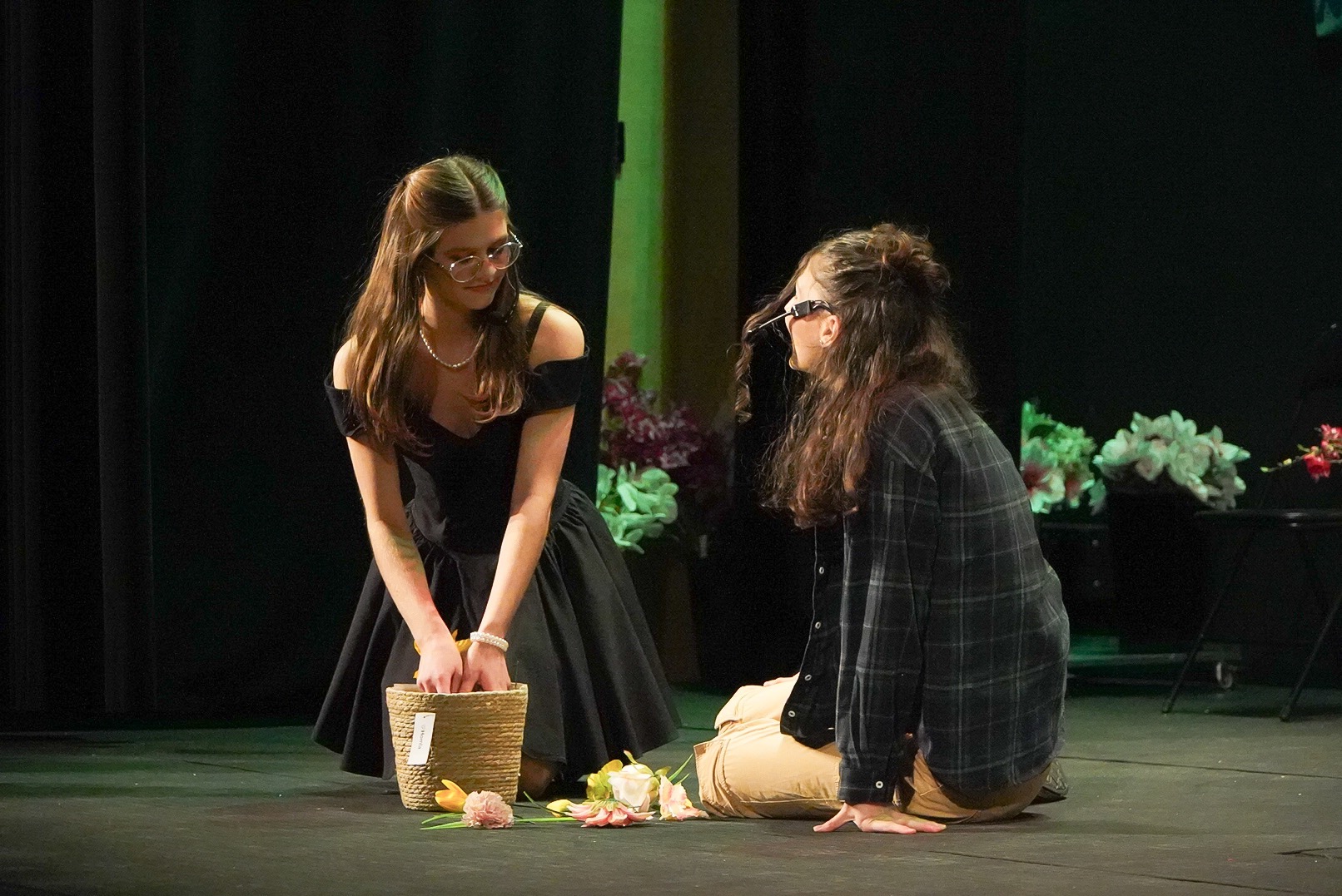 Dwie kobiety na scenie, jedna w czarnej sukience z koszykiem i kwiatami, druga w kraciastej koszuli, oświetlenie sceniczne