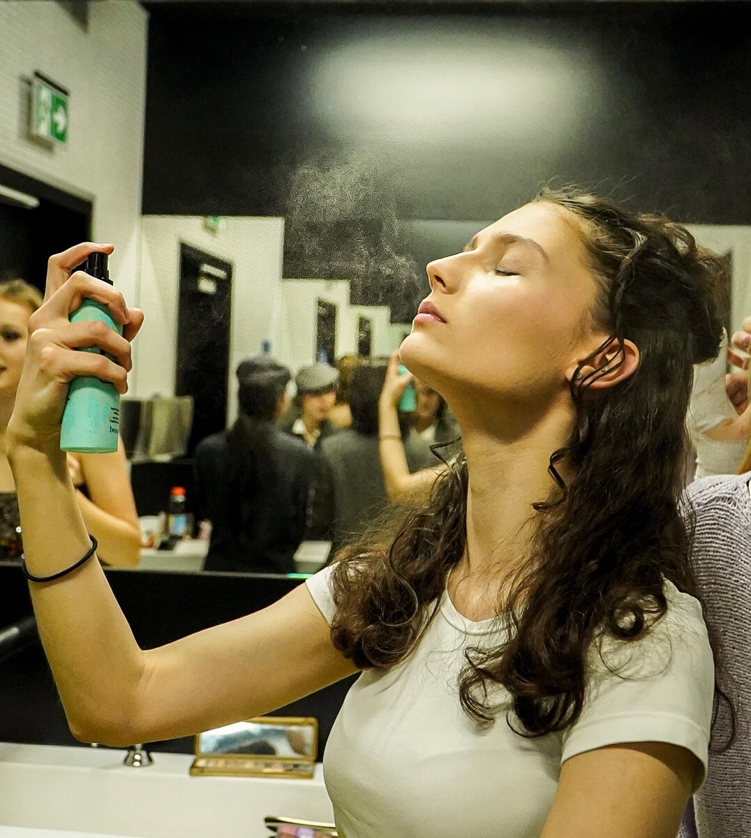 Kobieta z zamkniętymi oczami, spryskiwana mgiełką z butelki w pomieszczeniu z lustrami i innymi osobami w tle, prawdopodobnie podczas przygotowań do sesji zdjęciowej.