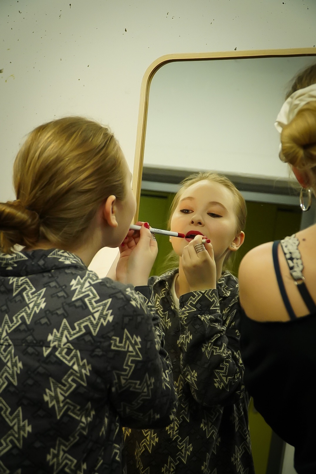 Młoda kobieta nakłada czerwoną szminkę przed lustrem w drewnianej ramie, widoczne odbicie i część drugiej osoby z tyłu.