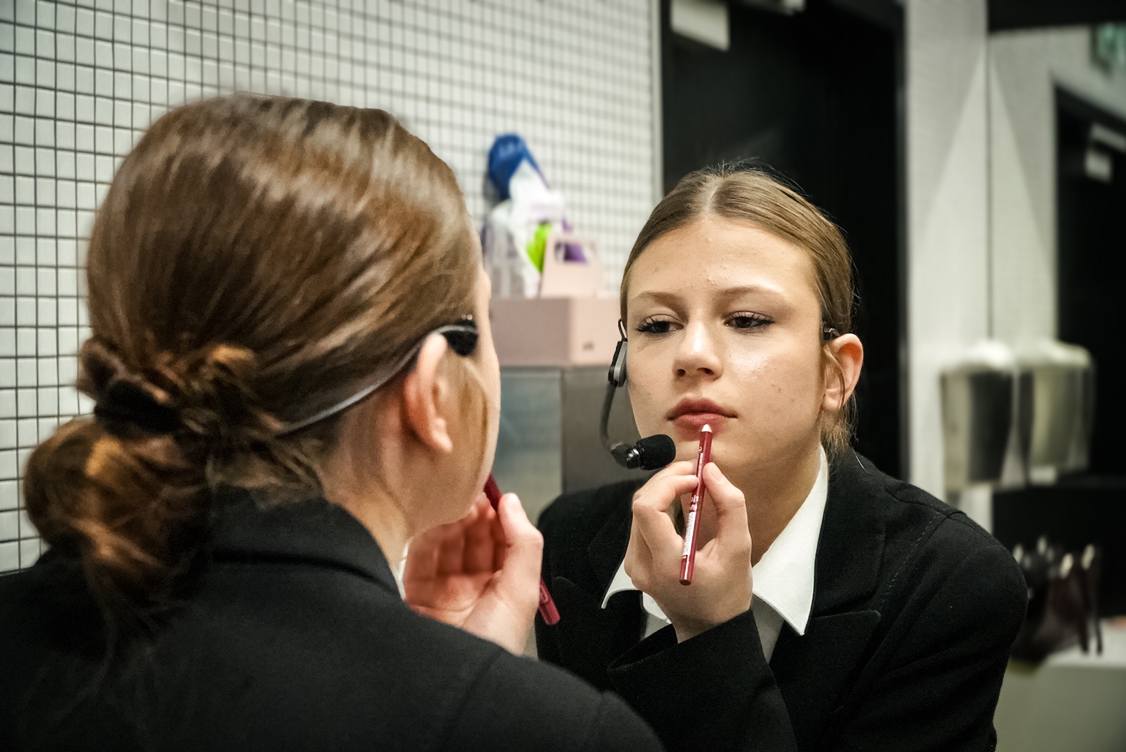 Młoda kobieta w czarnym garniturze, z mikrofonem przy uchu, maluje usta czerwoną kredką przed lustrem w pomieszczeniu z białą, kwadratową mozaiką na ścianach.