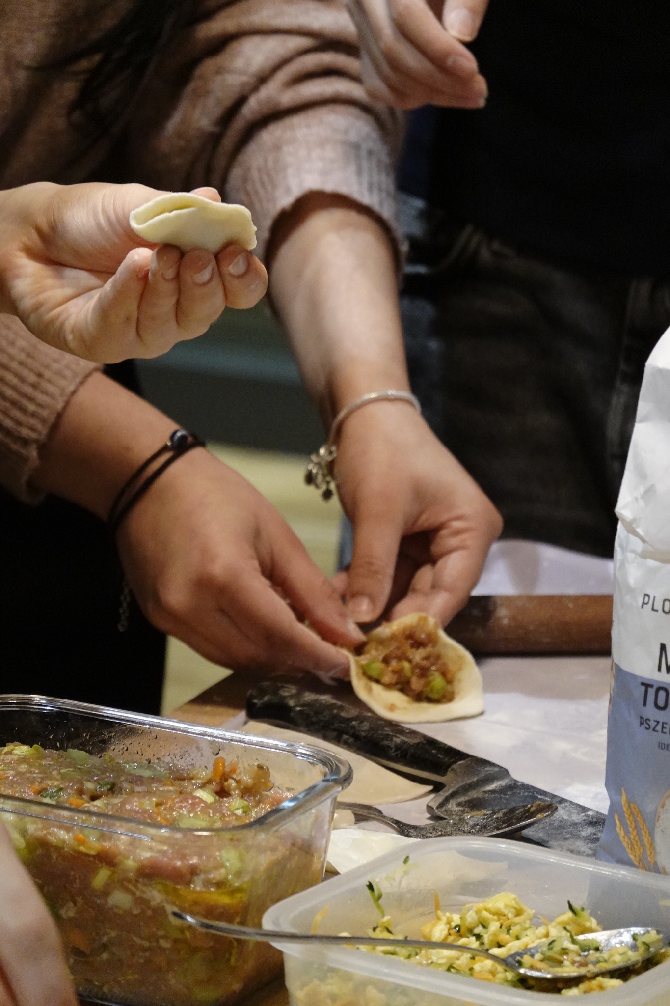 Przygotowywanie pierogów: dłonie formują ciasto z farszem, widoczne składniki w pojemnikach, wałek i mąka na stole.