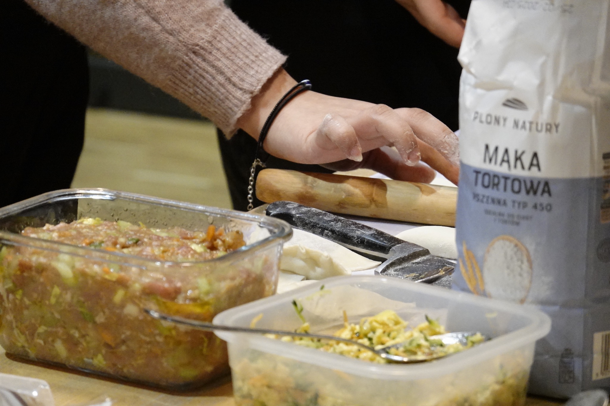 Przygotowywanie farszu i ciasta na pierogi: dłoń posypana mąką, wałek, nóż, dwa pojemniki z farszem i paczka mąki tortowej.