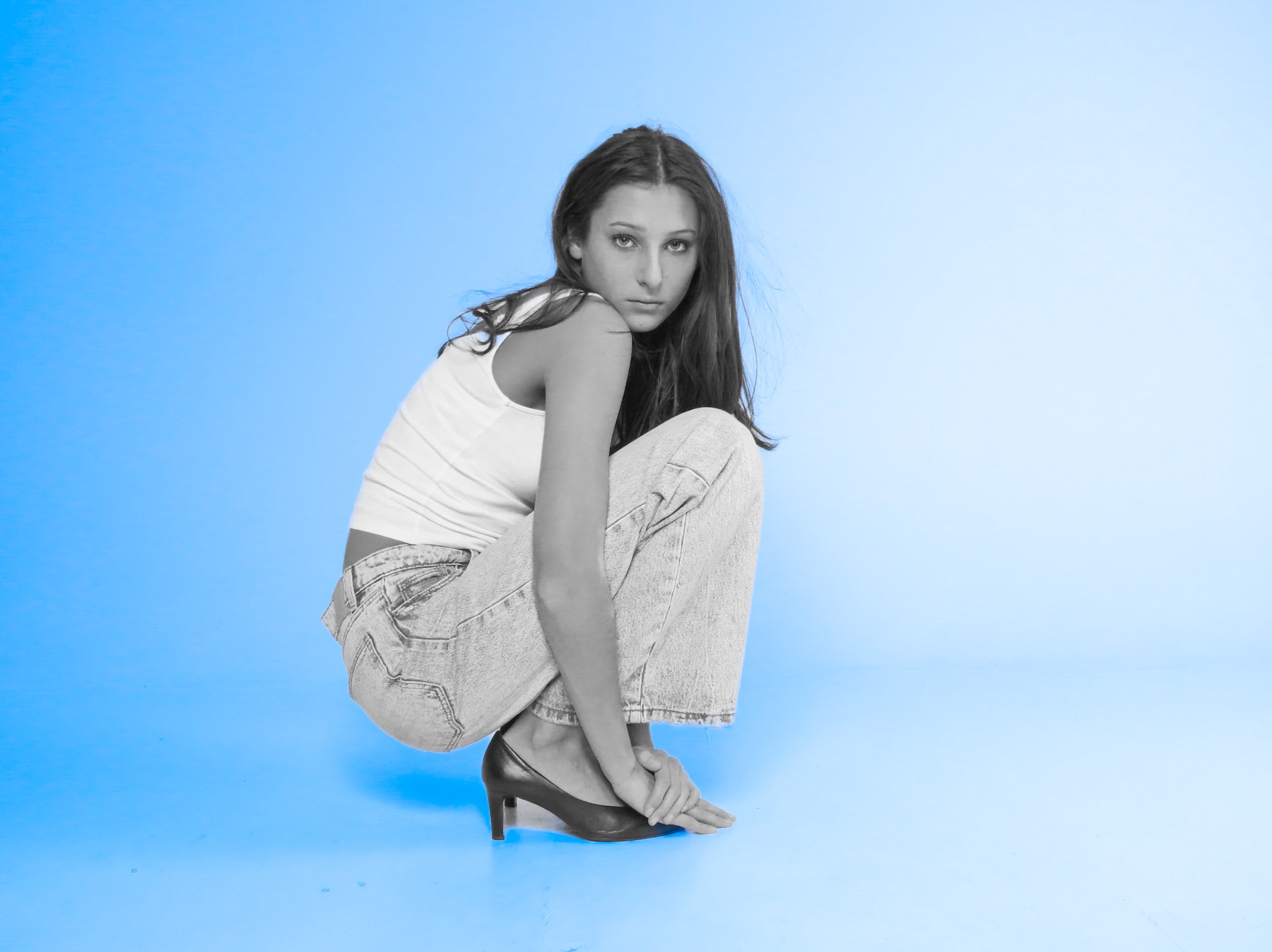 Kobieta w białym topie i jeansach w kucki, z rękami na stopie w czarnym szpilce, na jednolitym błękitnym tle.
