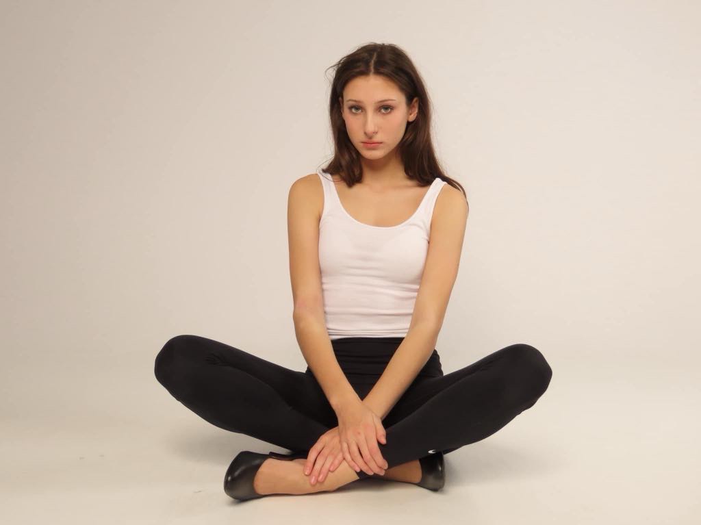 Kobieta siedząca po turecku na podłodze w białym topie i czarnych legginsach, dłonie splecione na kolanach, neutralny wyraz twarzy, jednolite jasne tło.