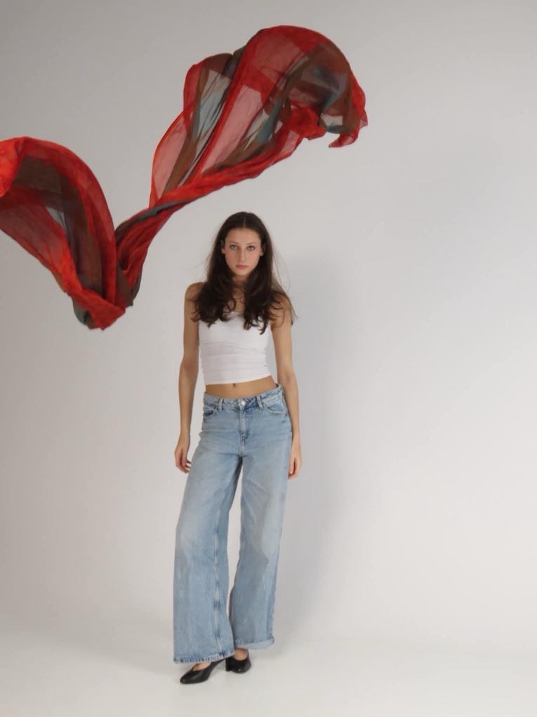 Młoda kobieta w białym topie i szerokich jeansach pozuje na białym tle z powiewającą, półprzezroczystą chustą w odcieniach czerwieni i zieleni.