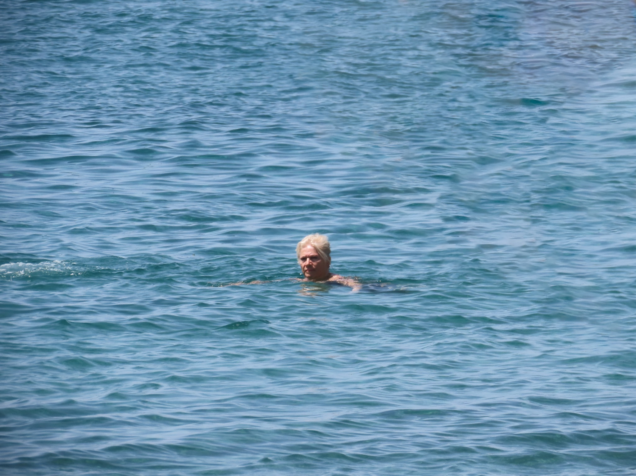 Kobieta z blond włosami płynie w błękitnej, lekko falującej wodzie, widoczna od ramion w górę.