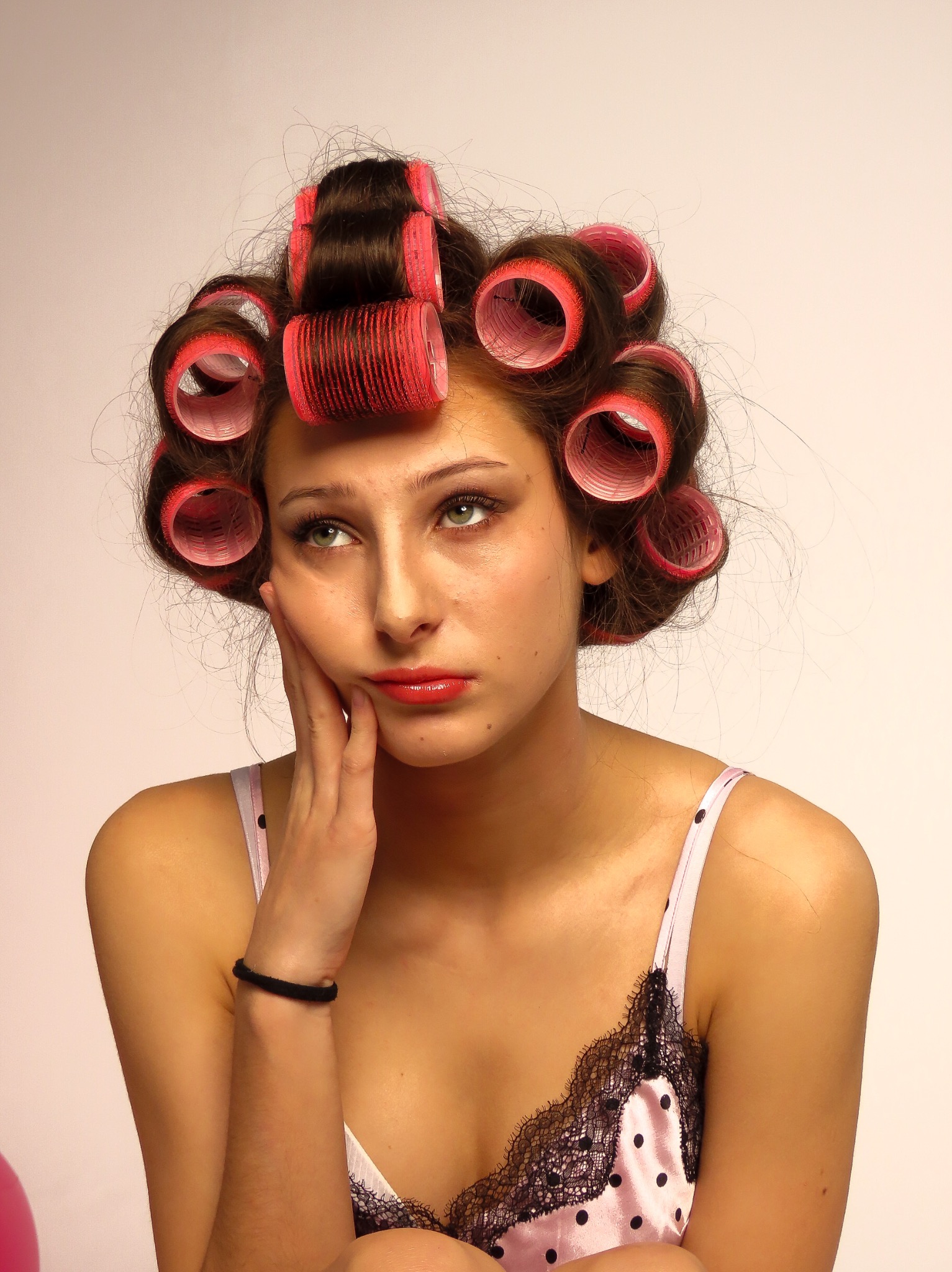 Portret kobiety z czerwonymi wałkami na włosach, opierającej brodę na dłoni, w satynowej koszulce nocnej w groszki z koronkowym wykończeniem.