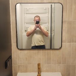 PR Wykończenia - Beżowa łazienka po remoncie z umywalką, złotą baterią i lustrem, w którym odbija się osoba robiąca zdjęcie. Na ścianie dekoracyjna lampa.