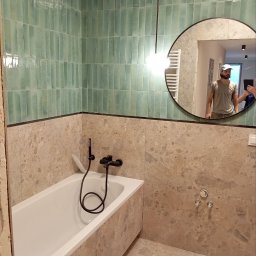 PR Wykończenia - Wykończona łazienka z wanną obłożoną marmurem, turkusowymi płytkami na ścianie i okrągłym lustrem, w którym odbija się osoba.