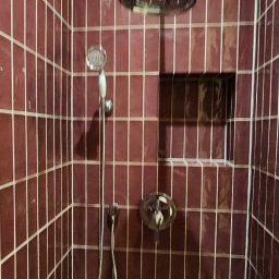 PR Wykończenia - Wykończona kabina prysznicowa z bordowymi, błyszczącymi płytkami, z wnęką na kosmetyki i deszczownicą. Widoczna bateria i słuchawka prysznicowa.