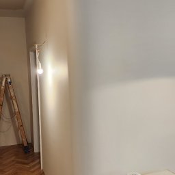 PR Wykończenia - Gładka, świeżo pomalowana ściana z zaokrąglonym narożnikiem, klimatyzator na górze, drabina malarska i kabel na parkiecie w jodełkę. Wnętrze po remoncie.