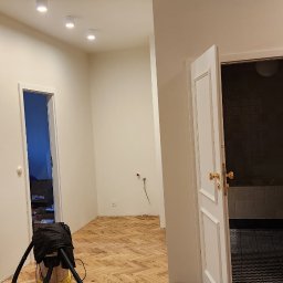 PR Wykończenia - Parkiet ułożony w jodełkę po renowacji, widoczny odkurzacz przemysłowy, białe ściany i otwarte drzwi do łazienki. Całość oświetlona ciepłym światłem.