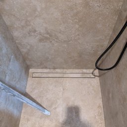 PR Wykończenia - Kabina prysznicowa z beżowymi płytkami i odpływem liniowym. W rogu widoczny fragment buta i element kabiny.