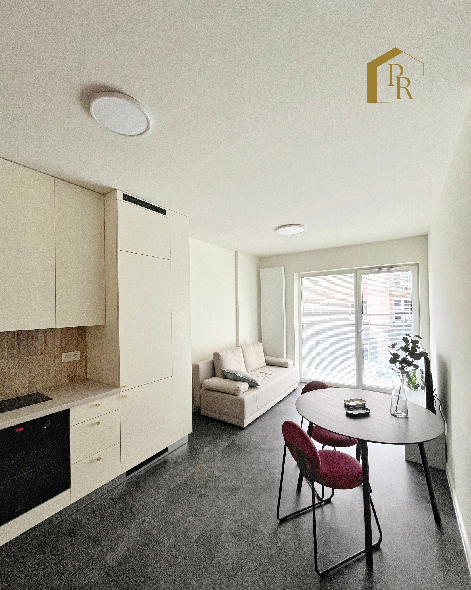 Jasne wnętrze mieszkania po remoncie: minimalistyczna kuchnia, stół z krzesłami i sofa przy oknie. Nowoczesne oświetlenie i ciemna podłoga.