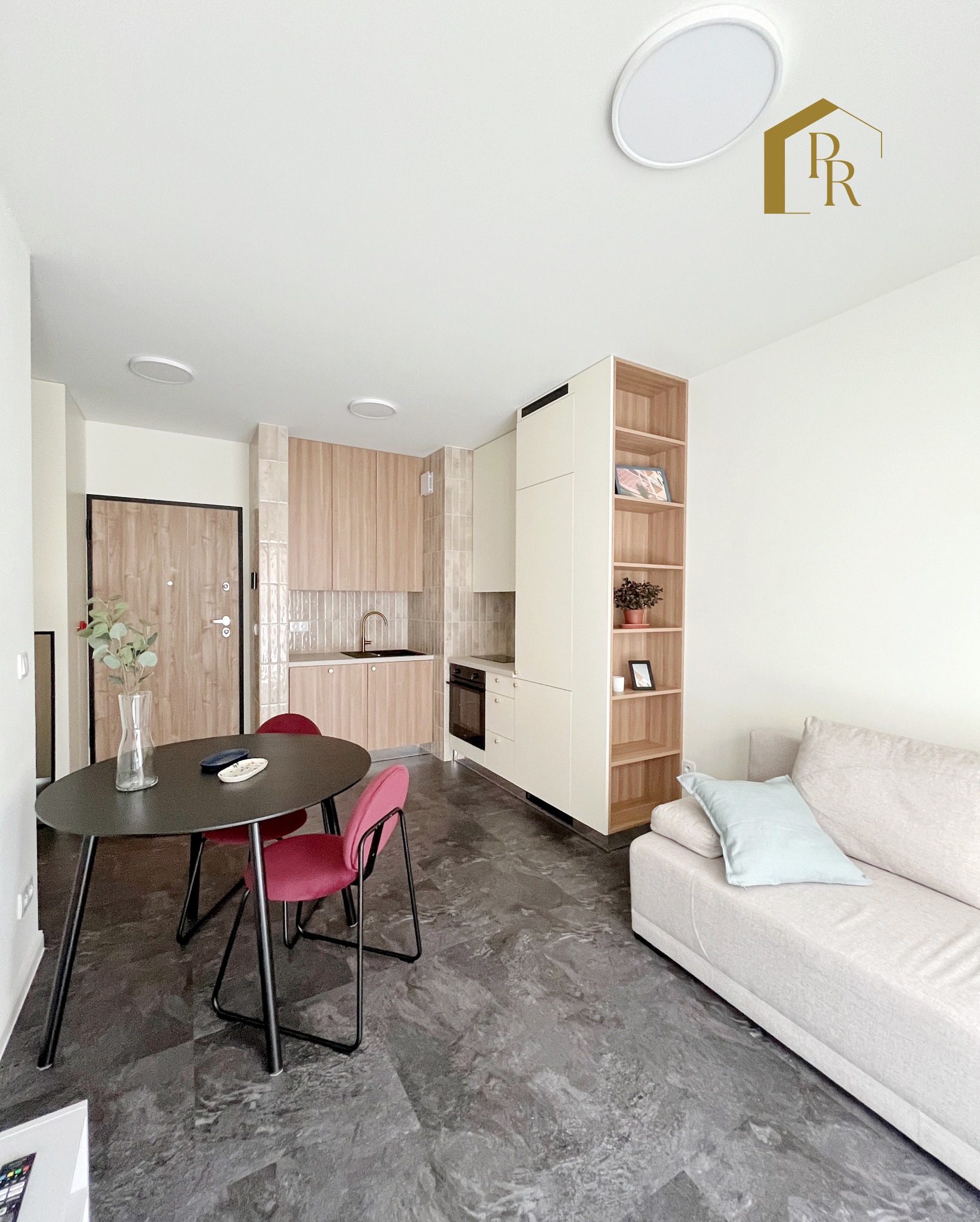 Jasne wnętrze mieszkania po remoncie: stół z krzesłami, sofa, zabudowa kuchenna z drewnianymi frontami i regał. Podłoga w odcieniach szarości imitująca kamień.