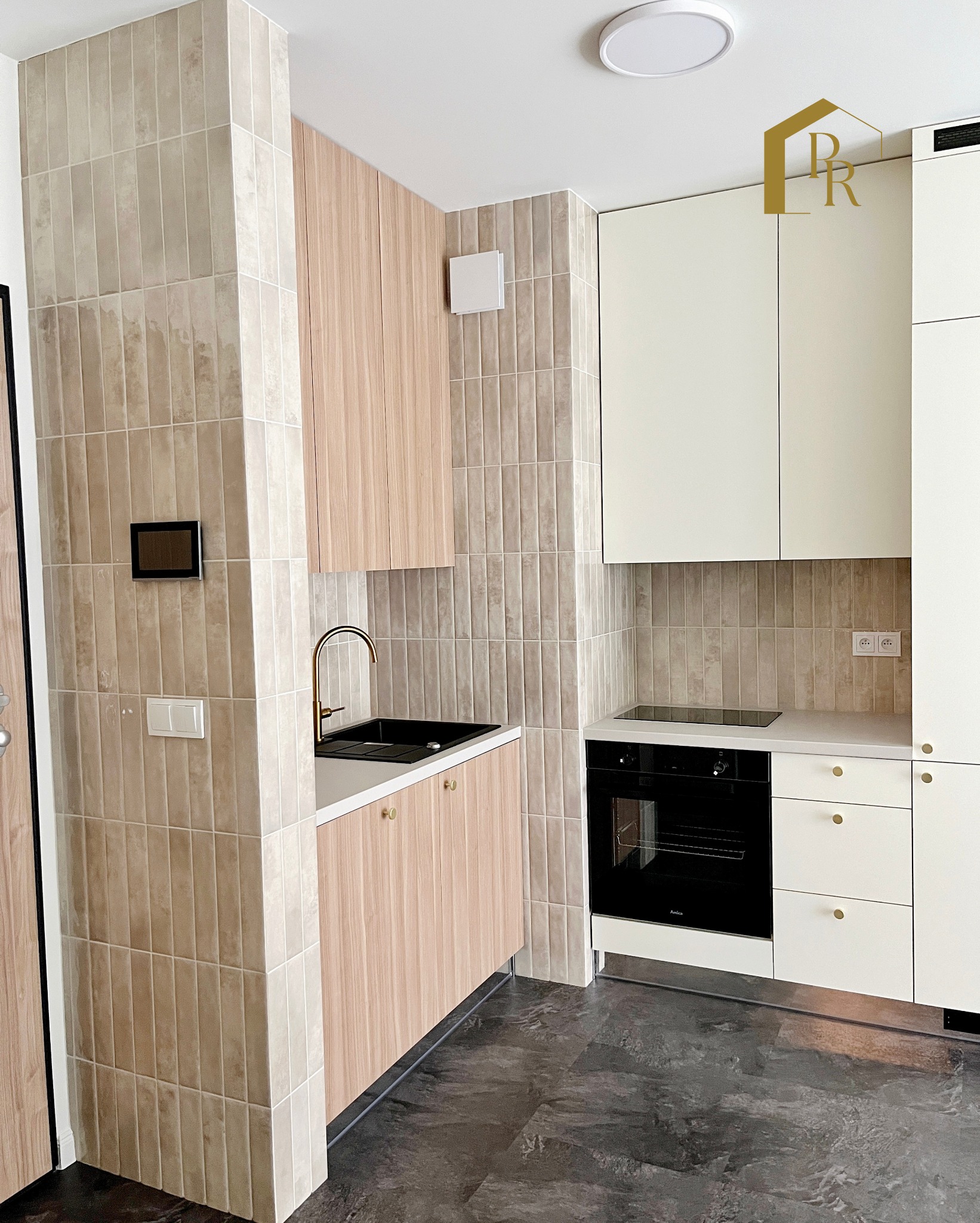 Nowoczesna kuchnia z beżowymi płytkami ułożonymi pionowo, drewnianymi szafkami i czarnym zlewem z złotą armaturą. Widoczny fragment podłogi z ciemnego gresu.