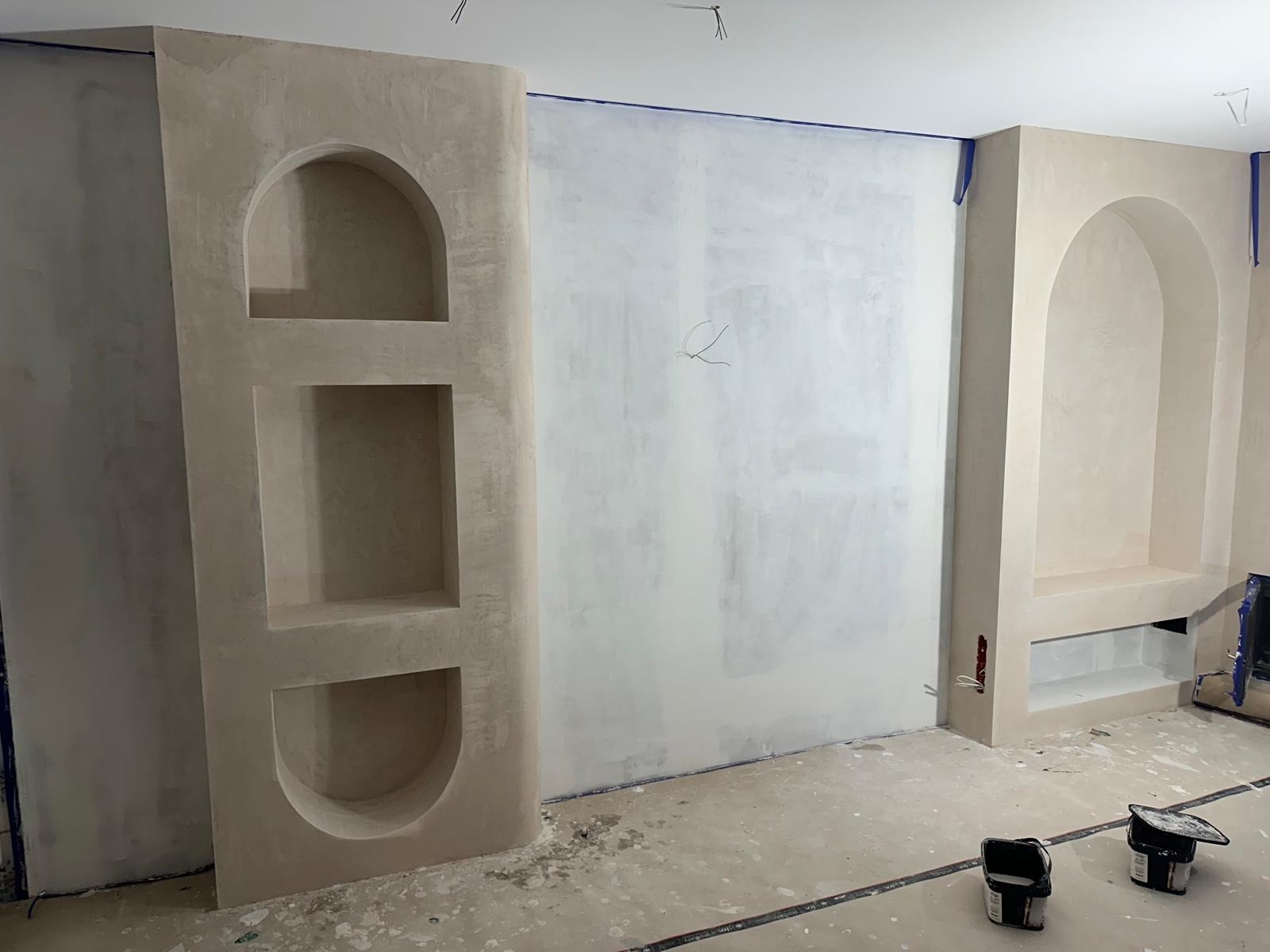 Wnętrze w trakcie prac wykończeniowych: ściany po szpachlowaniu z wnękami o nieregularnych kształtach. Widoczne narzędzia malarskie i taśmy zabezpieczające.