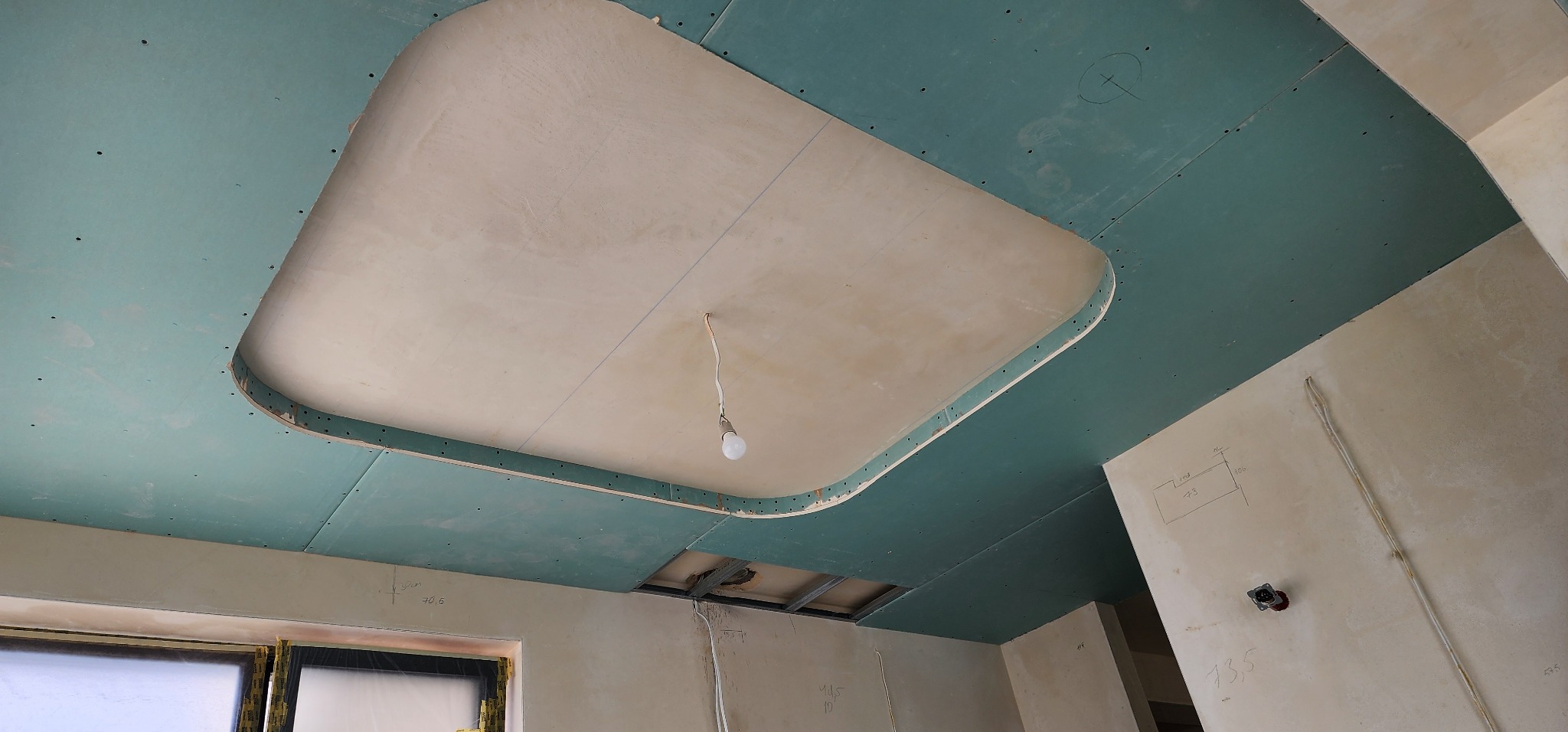 Sufit podwieszany z płyt gipsowo-kartonowych w trakcie montażu. Widoczne profile i oświetlenie tymczasowe. Ściany w stanie surowym z naniesionymi oznaczeniami.