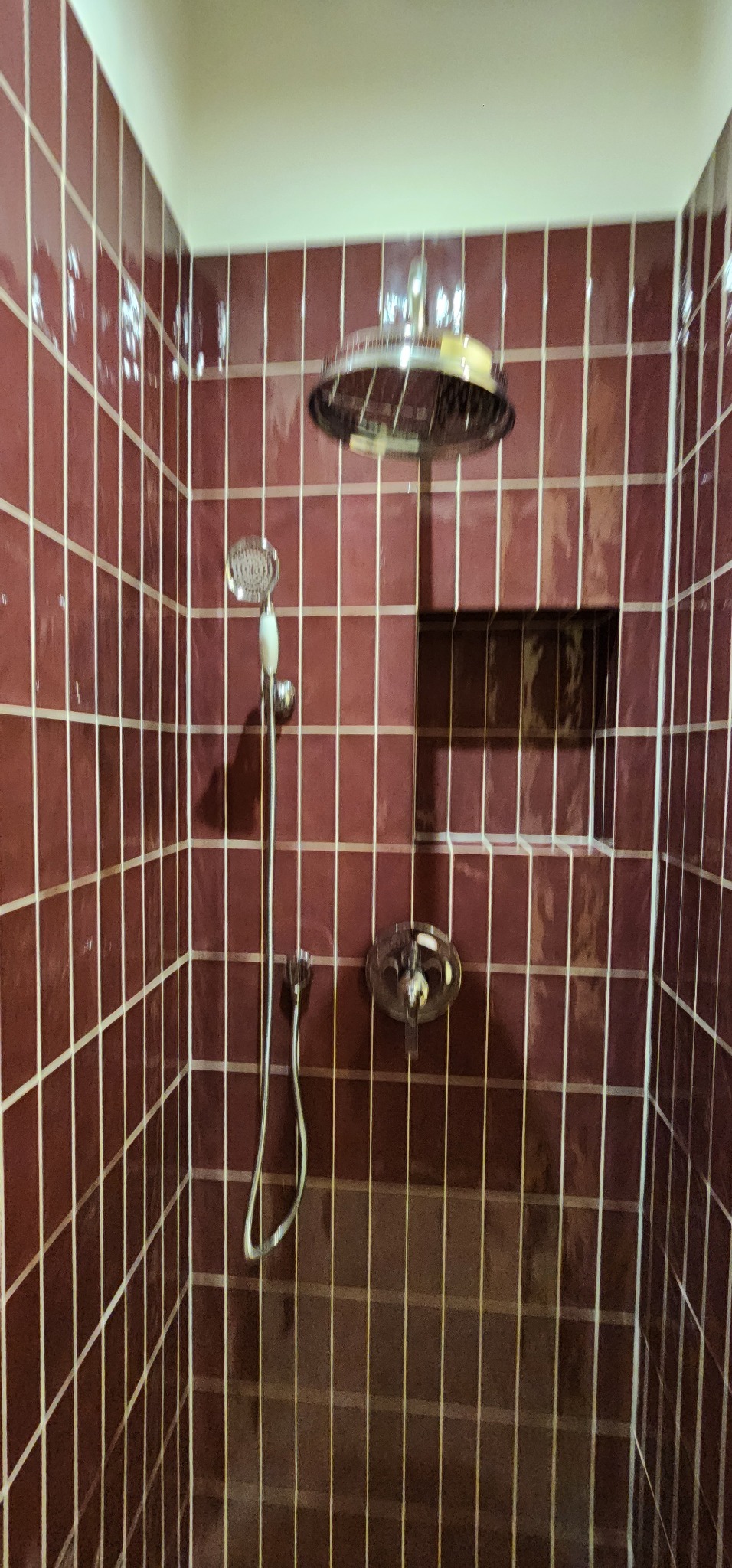 Wykończona kabina prysznicowa z bordowymi, błyszczącymi płytkami, z wnęką na kosmetyki i deszczownicą. Widoczna bateria i słuchawka prysznicowa.