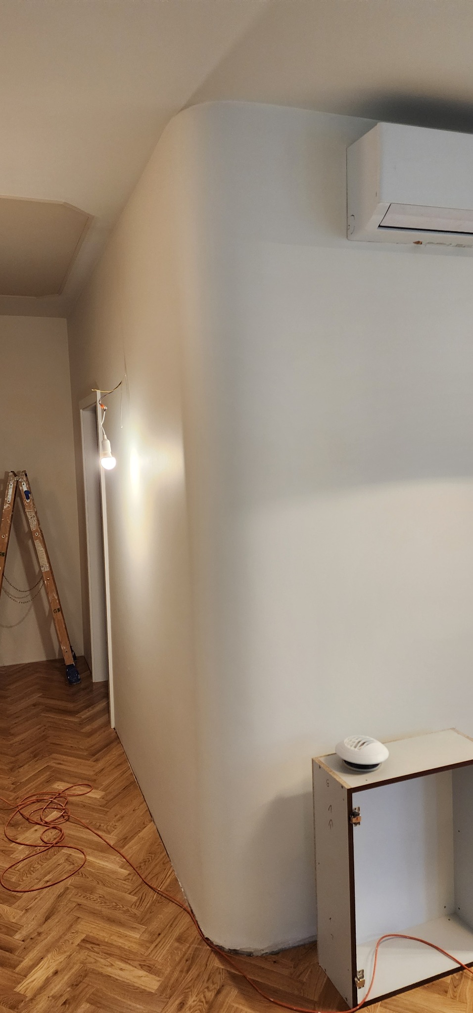 Gładka, świeżo pomalowana ściana z zaokrąglonym narożnikiem, klimatyzator na górze, drabina malarska i kabel na parkiecie w jodełkę. Wnętrze po remoncie.