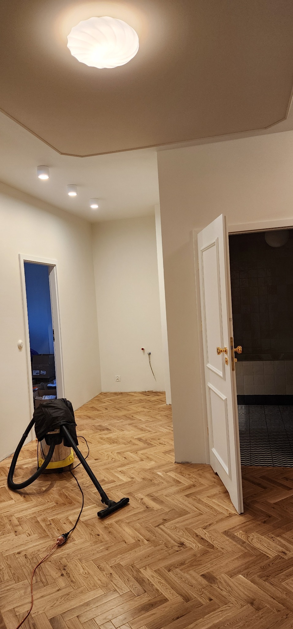 Parkiet ułożony w jodełkę po renowacji, widoczny odkurzacz przemysłowy, białe ściany i otwarte drzwi do łazienki. Całość oświetlona ciepłym światłem.