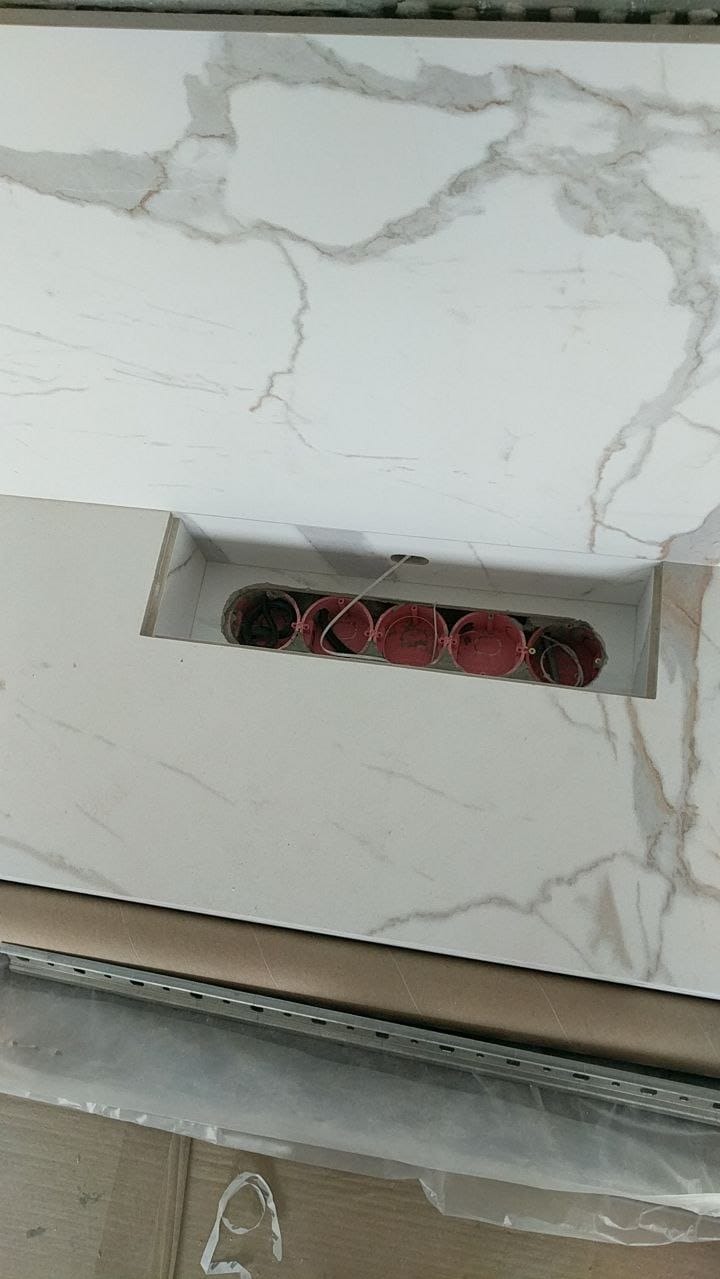 Marmurowe płytki z wyciętym otworem na gniazdka elektryczne, widoczne czerwone puszki instalacyjne. Etap prac wykończeniowych, oświetlenie sufitowe.