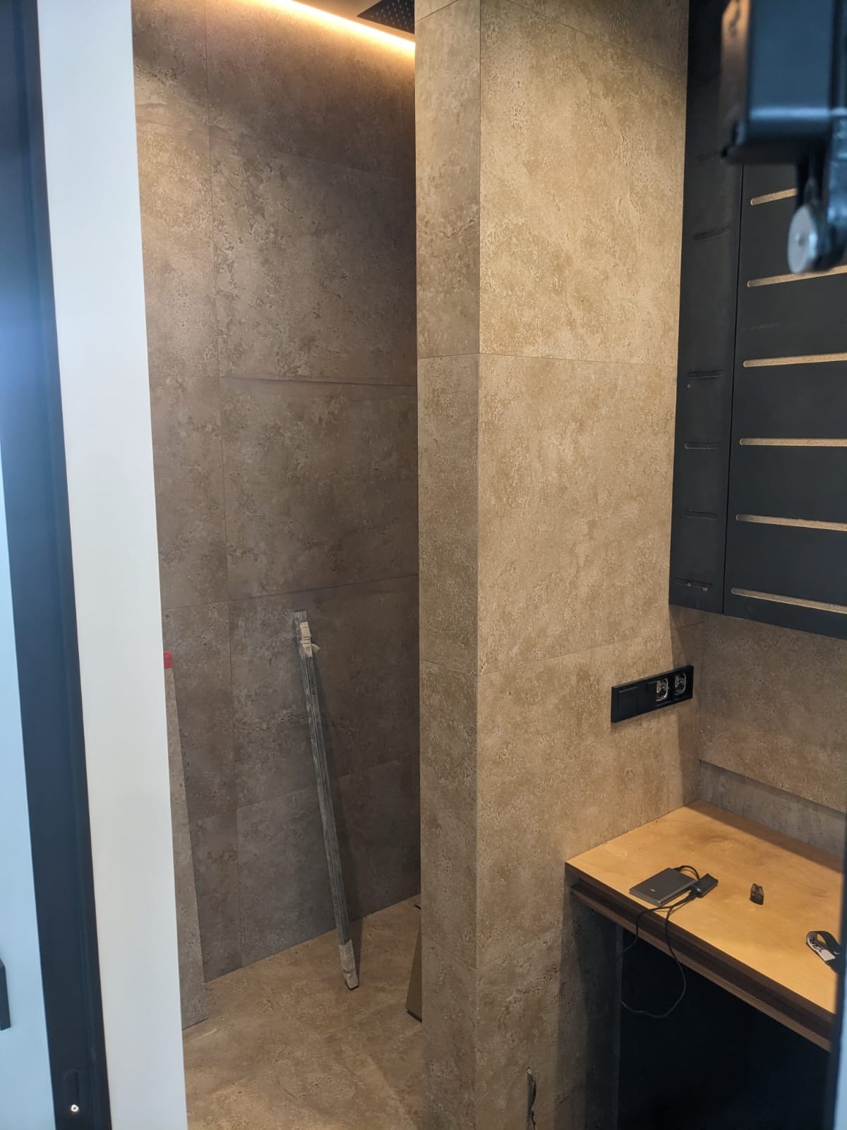 Beżowa łazienka w trakcie wykańczania: płytki imitujące kamień, czarne gniazdka i szafka, listwa przypodłogowa oparta o ścianę. Oświetlenie sufitowe.