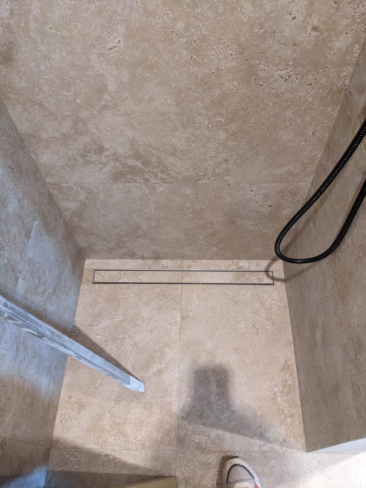 Kabina prysznicowa z beżowymi płytkami i odpływem liniowym. W rogu widoczny fragment buta i element kabiny.