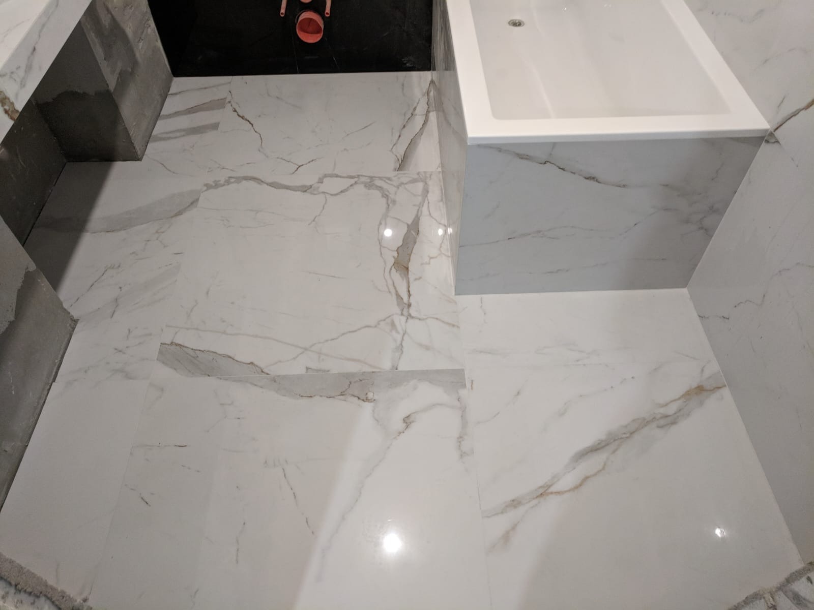 Białe, marmurowe płytki w łazience, ułożone na podłodze i ścianach wokół wanny. Widoczne fragmenty niedokończonych prac przy zabudowie umywalki.