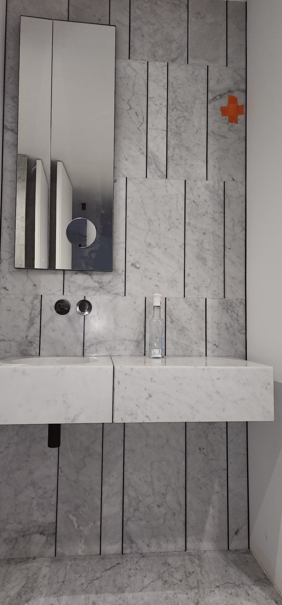 Nowoczesna łazienka z marmurową umywalką, lustrem i minimalistyczną armaturą. Na ścianie widoczny pomarańczowy znak. Estetyczne wykończenie wnętrza.