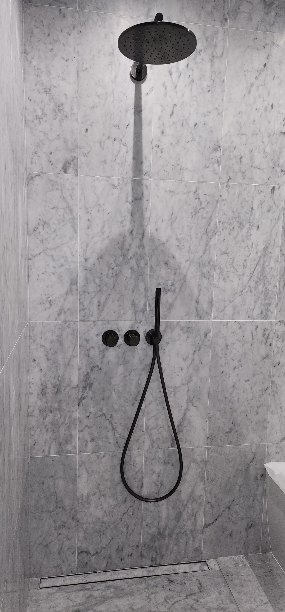 Nowoczesna kabina prysznicowa z czarną armaturą na tle marmurowych płytek. Minimalistyczny design z odpływem liniowym.