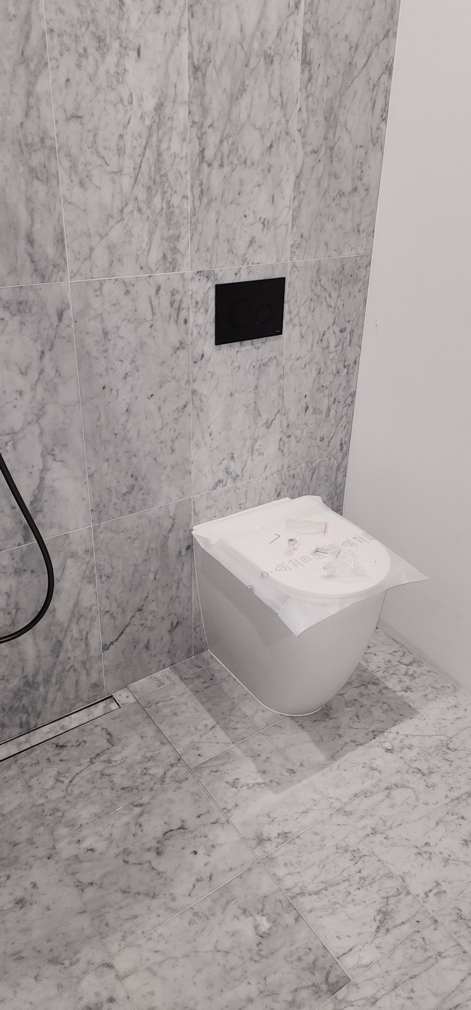 Nowoczesna toaleta podtynkowa, jeszcze w folii ochronnej, zamontowana na ścianie wyłożonej marmurowymi płytkami. Widoczny czarny przycisk spłukujący.